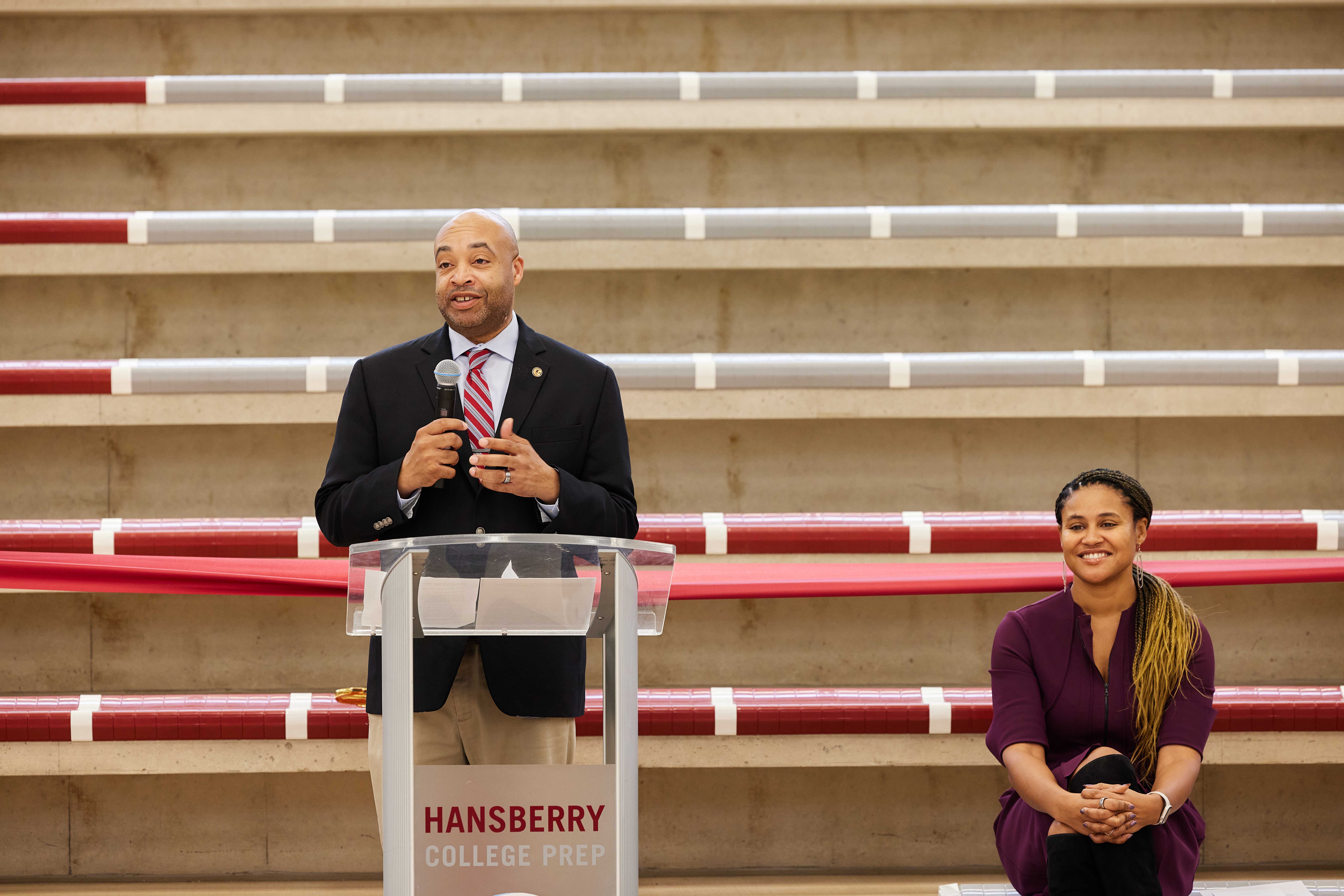 This photo shows Illinois State Representative Justin Slaughter speaking behind a podium at the Hansberry College Prep new gym renovation ribbon-cutting ceremony. Behind him, Noble Schools' CEO Constance Jones is sitting on the brand new bleachers.