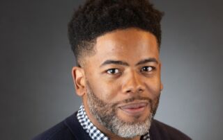 This photo shows a professional headshot of Justin Boyd, the music teacher and band director at UIC College Prep. He is a Black man with short black hair and a gray and black beard. He is wearing a blue and white checkered button-up with a dark blue sweater over it.