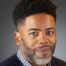 This photo shows a professional headshot of Justin Boyd, the music teacher and band director at UIC College Prep. He is a Black man with short black hair and a gray and black beard. He is wearing a blue and white checkered button-up with a dark blue sweater over it.