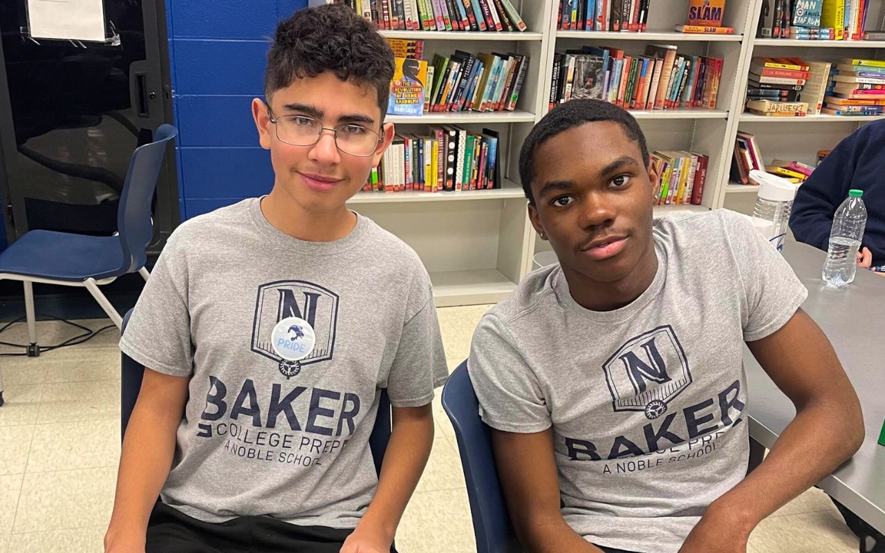 In this photo, two Baker College Prep students are sitting side by side and smiling at the camera. They are a mentor and mentee pair in the Lion's Pride Mentoring program during the 2023-2024 school year.