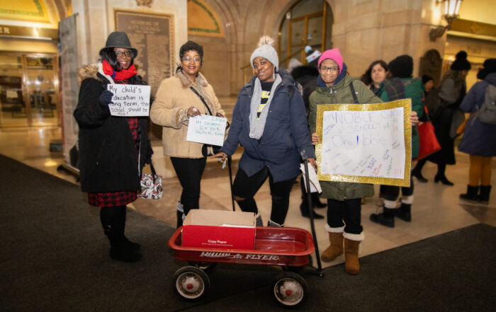 Noble parents stand in City Hall with a wagon full of flyers at their feet and signs in their hands. All their signs say different things to promote charter schools.