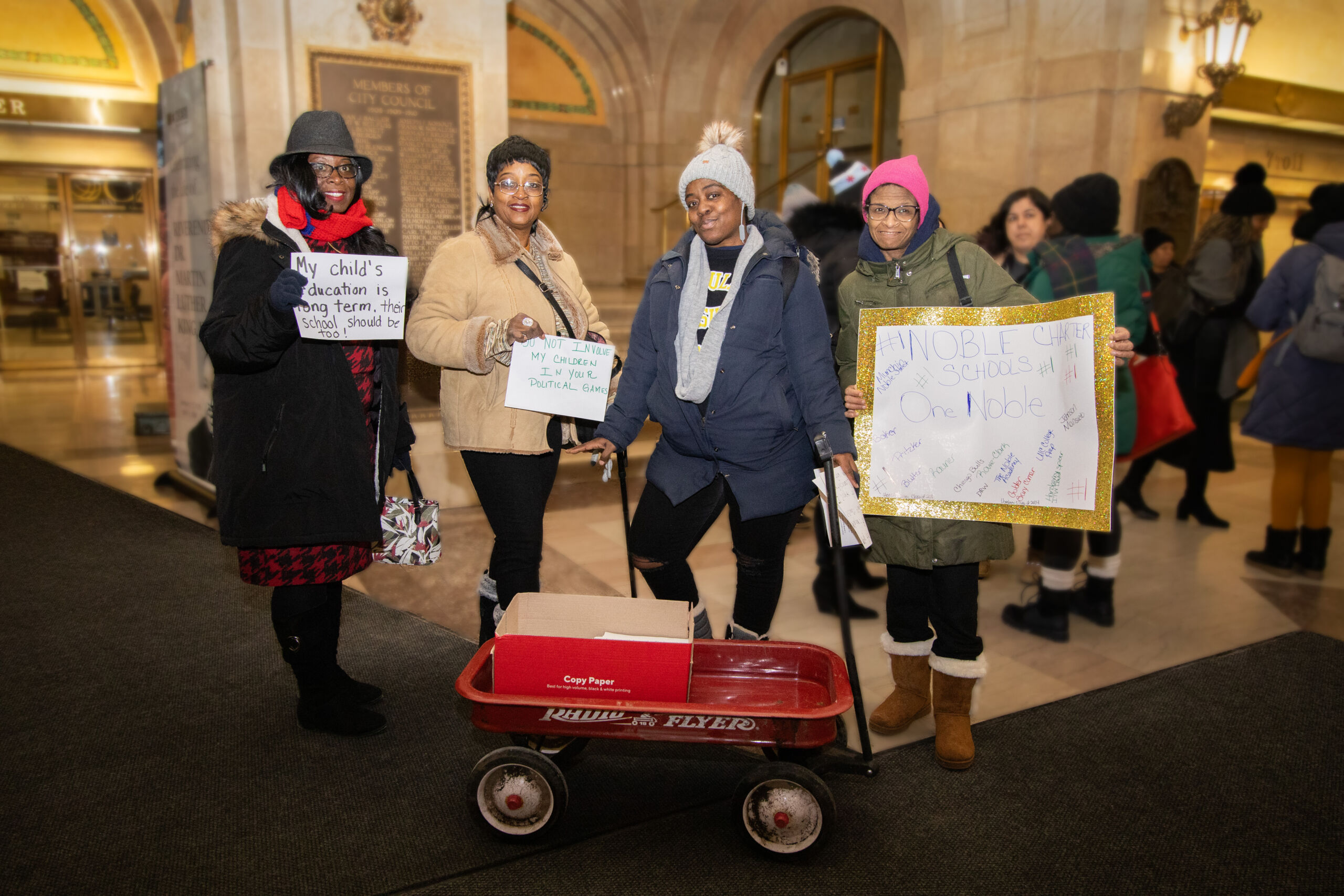 Noble parents stand in City Hall with a wagon full of flyers at their feet and signs in their hands. All their signs say different things to promote charter schools.