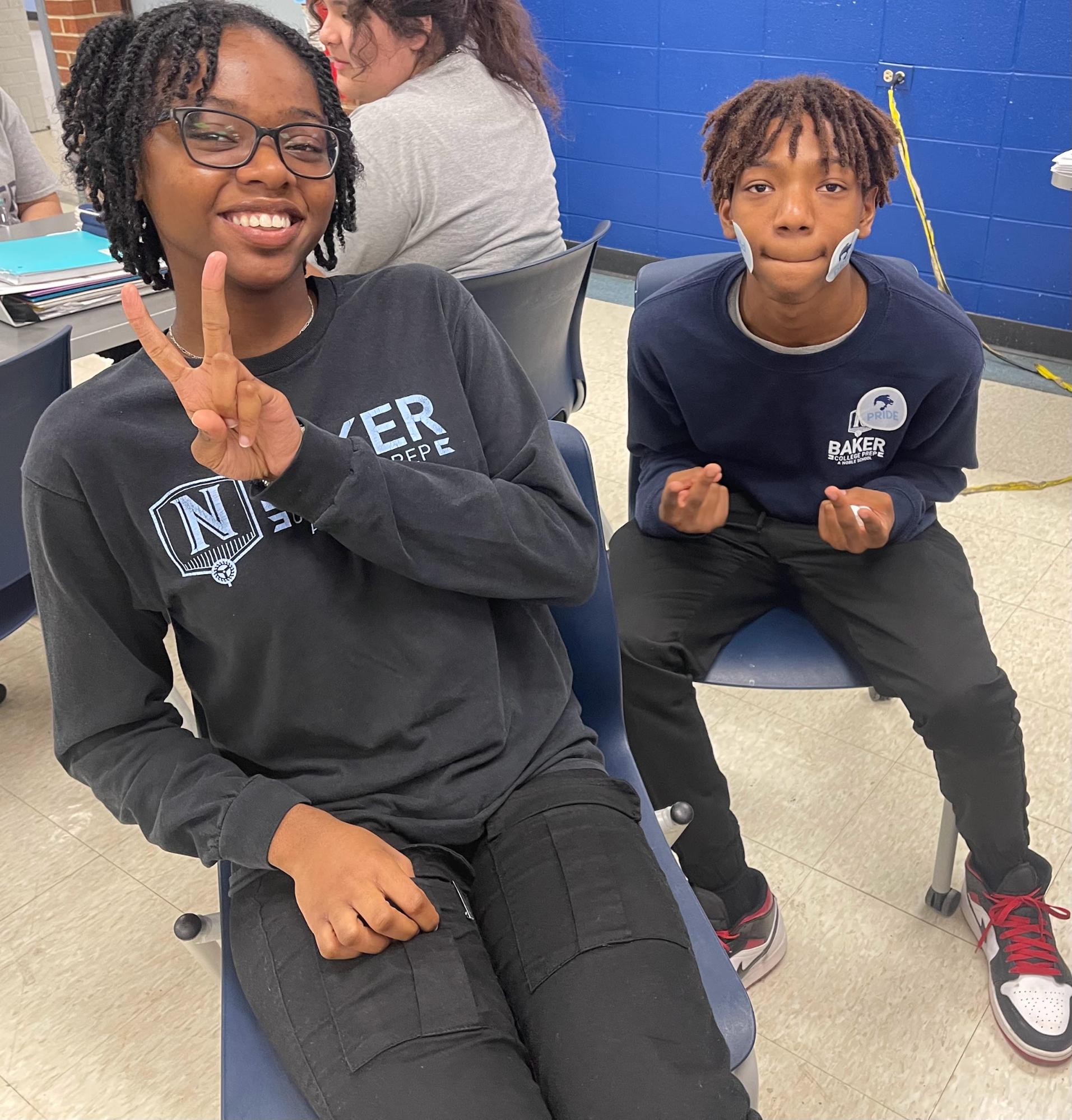 In this photo, Lion's Pride mentor and Baker College Prep junior Tykaha is sitting next to her mentee Tony. They are smiling and posing with peace signs for the camera.