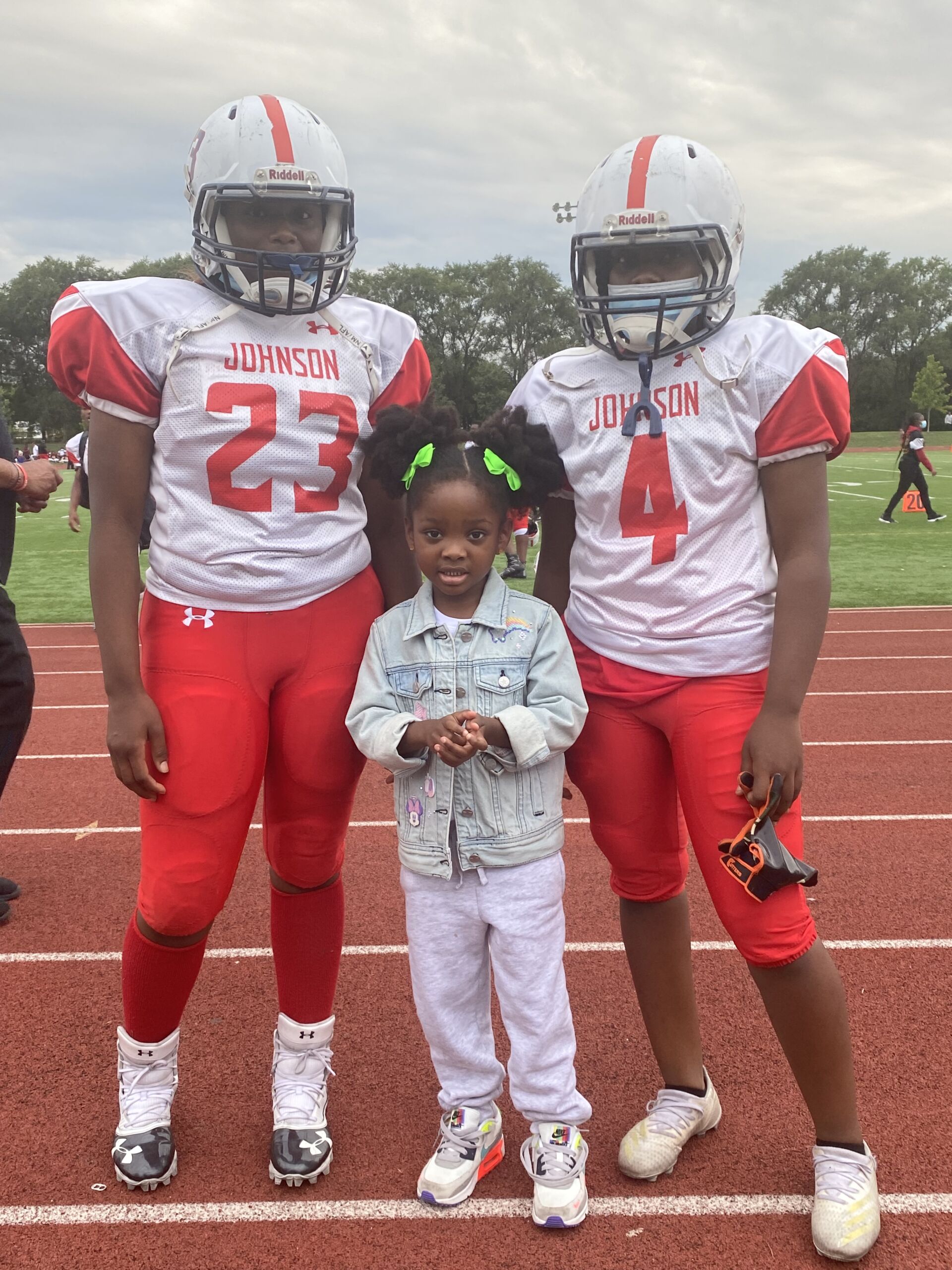 Image shows two female football players at Johnson College Prep, one of them Le'Andra H, standing with a young Black girl---Principal Jonas Cleaves' daughter. They are outside on the football field.
