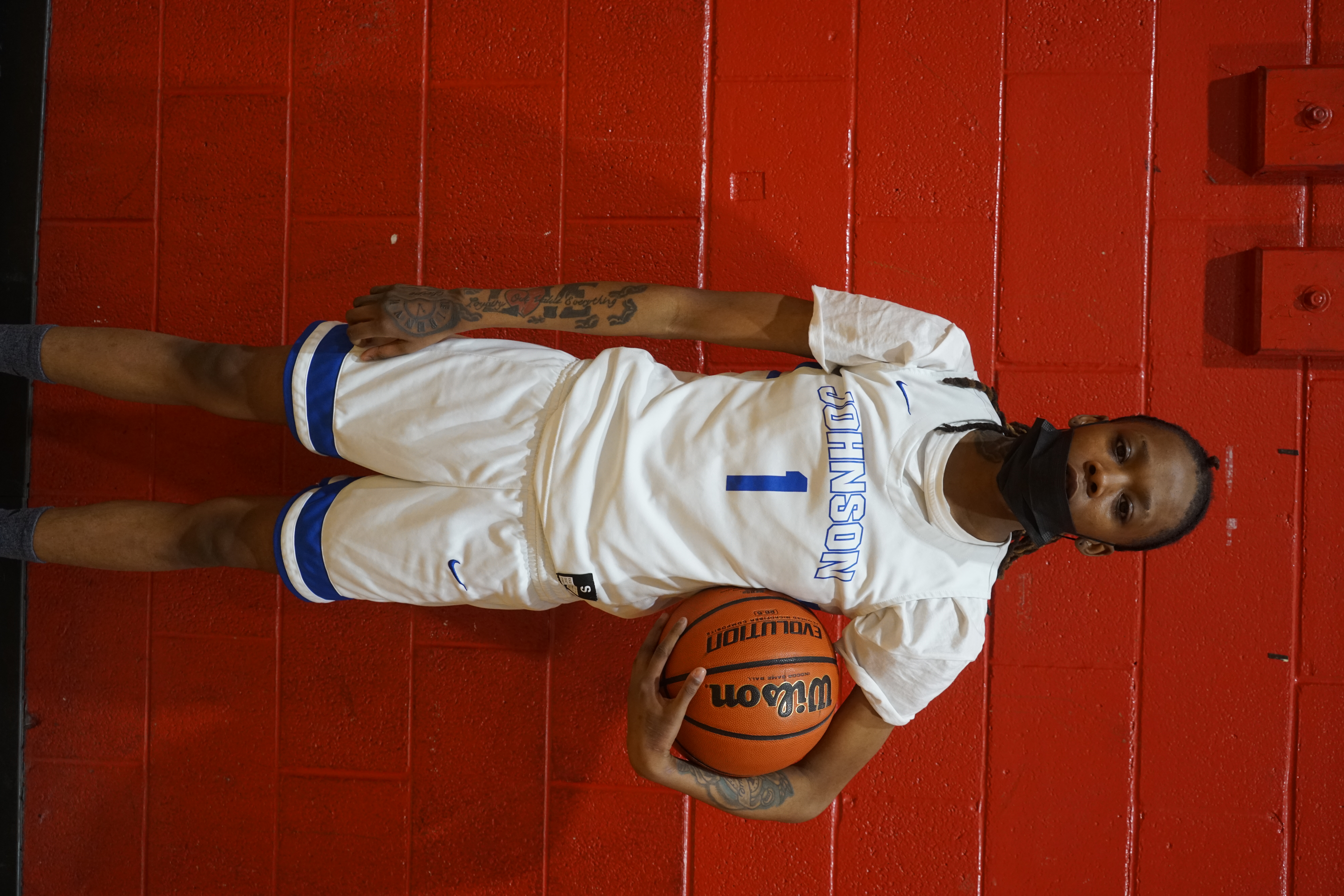 DSC06232 Photo is a full body portrait of Destiny T, a basketball player at Johnson College Prep. She is wearing a white and blue uniform. She has a basketball crooked in her left arm while her other arm falls to her side.