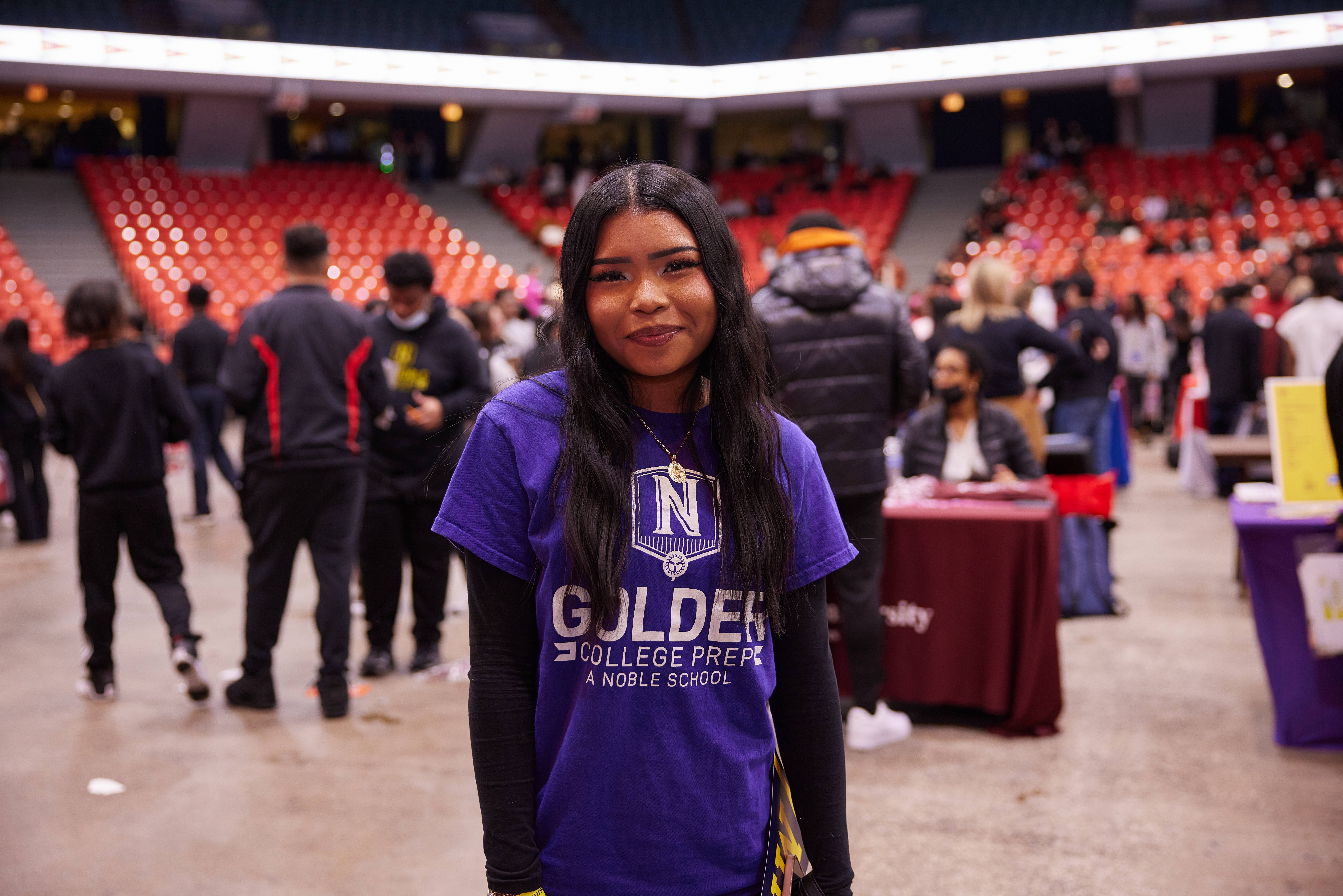 A Golder College Prep junior, Briana L, stands smiling in front of a bunch of college booths and crowd. She is at the Noble Schools' Junior College Fair and is wearing a bright purple Golder t-shirt.