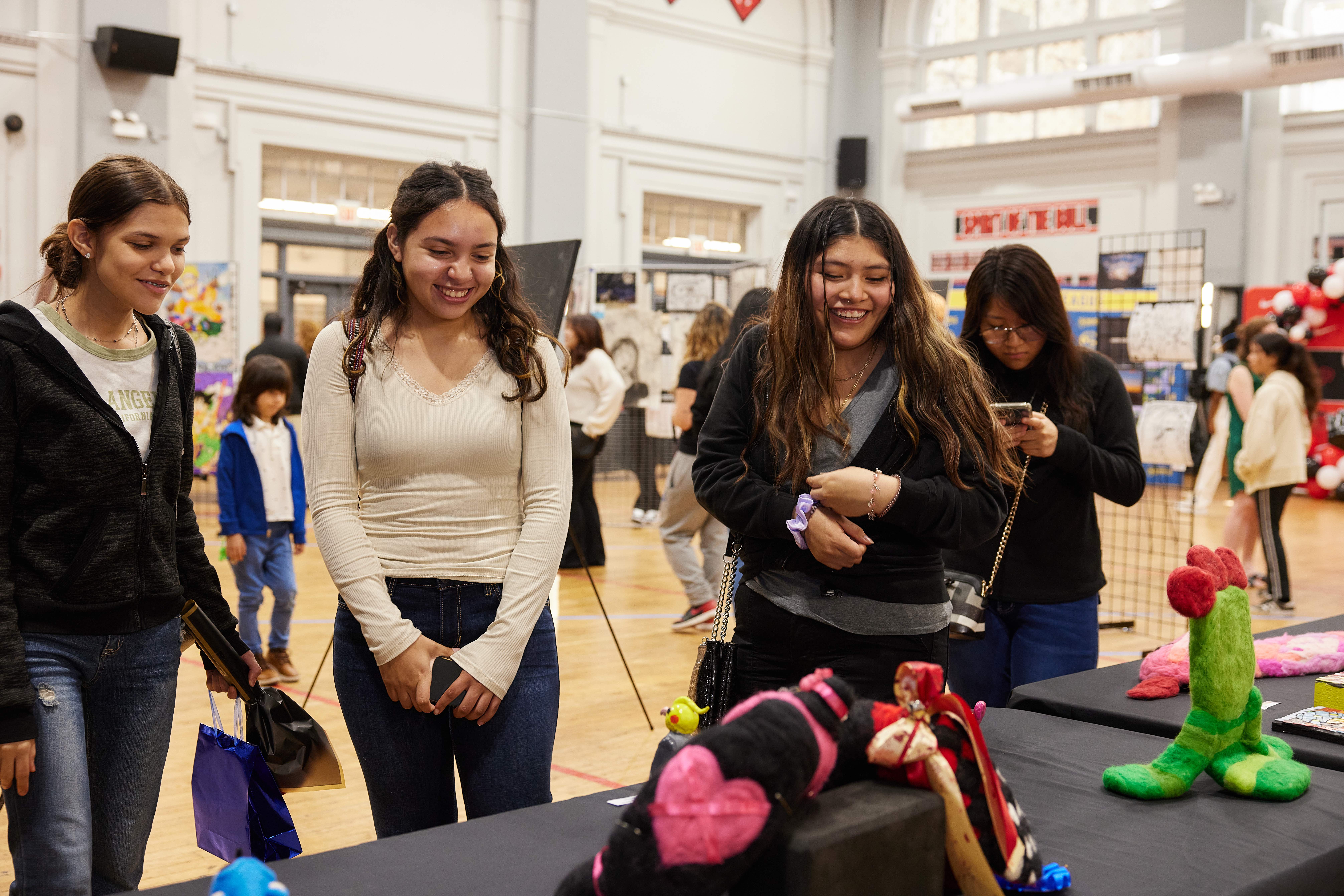 This image shows three Noble Schools students smiling at some art pieces on the table at the Visual Arts Festival. Behind them, you can see other people viewing all the student artwork displayed in the Chicago Bulls College Prep gym.