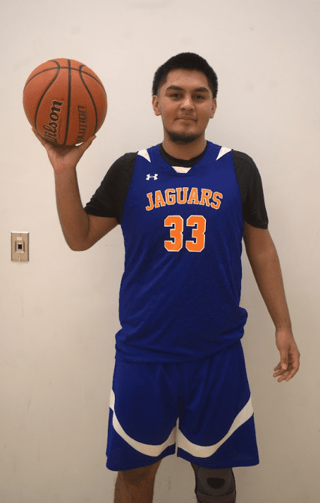 This photo is a full body shot of Adrian Zarco, a student at Pritzker College Prep. He is a young Latine man with short hair. In this photo, he is wearing his Pritzker Jaguar basketball uniform and is holding a basketball in one hand.