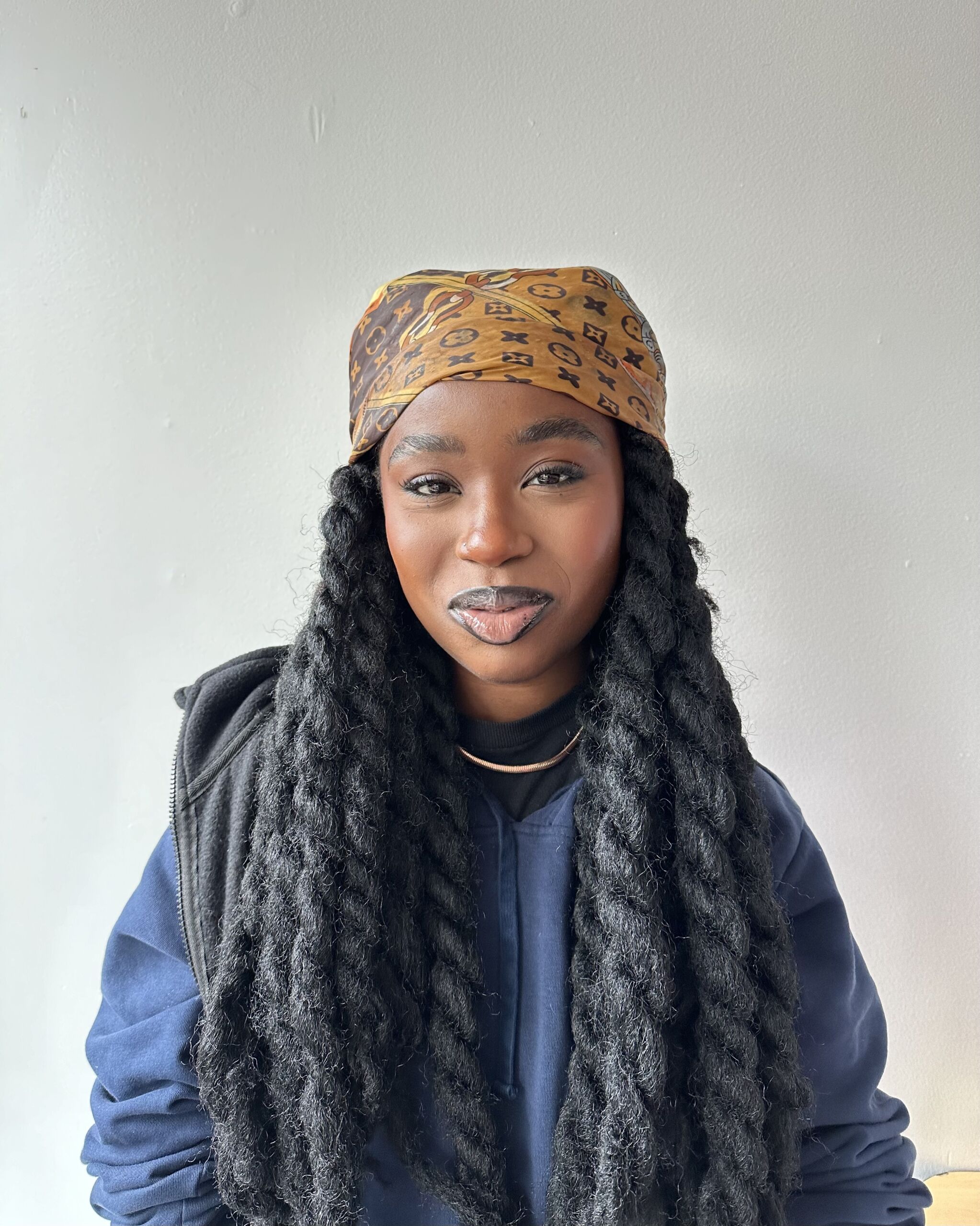 This photo is a headshot of Asiatu Mustapha, a student at The Noble Academy. She is a young Black woman with long hair in big braids. In this photo, she is smiling and wearing a patterned head scarf and a dark blue hoodie.