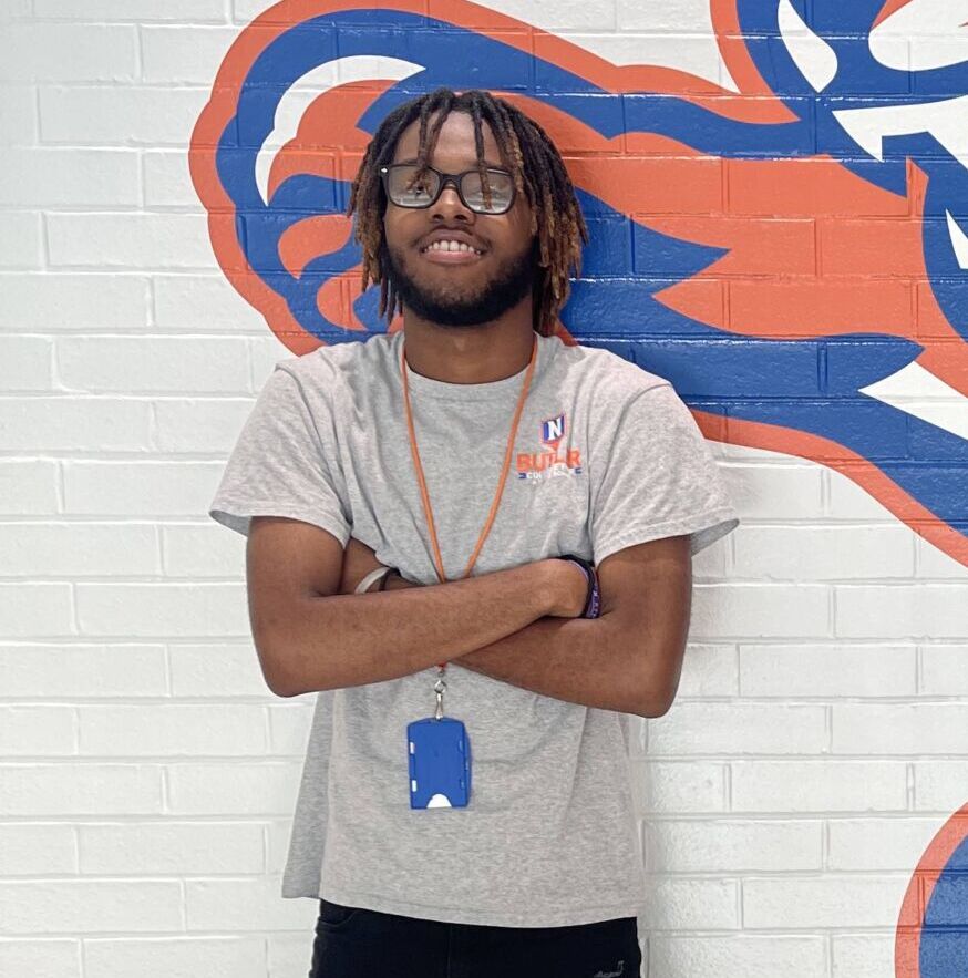 Photo shows a waist up shot of Damaris Purnell, a student at Butler College Prep. He is a young Black man with medium-length locs and glasses. He is smiling with his arms crossed and is wearing a light gray t-shirt with the Butler logo on it. Behind him, you can see a wall with the Butler mascot on it.