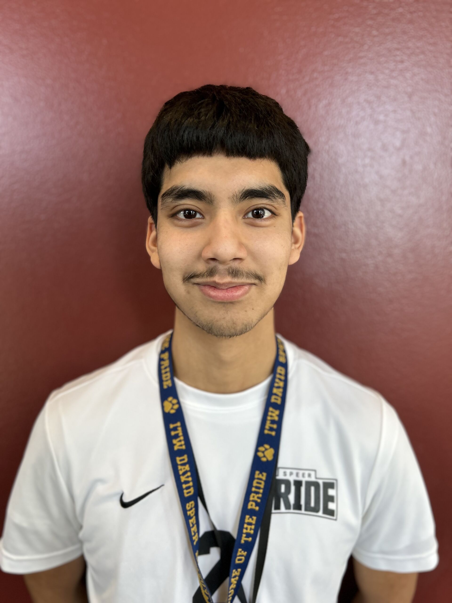 This photo shows a headshot of David Cano, a student at ITW David Speer Academy. He is a young Latine man with short hair. In this photo, he is smiling and wearing a white t-shirt with the Speer Pride logo on it.