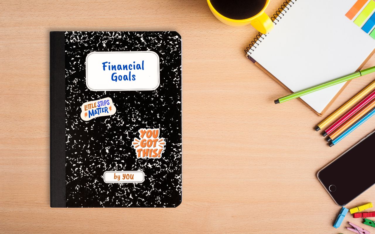 Photo shows a composition notebook on a wooden desk, surrounded by school supplies. On the composition notebook, it says "Financial Goals" in the title spot and "By YOU" in the author slot. There are two stickers on the notebook. One says "Little Steps Matter" and the other says "You Got This!"