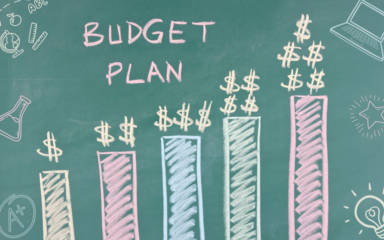 Image shows a chalkboard with a multicolored bar graph on it. The graph is labelled "Budget Plan" and each bar from lowest to highest has increasing dollar bill signs on top of them. On the edges of the chalkboard are education-themed doodles of pencils, laptops, and more