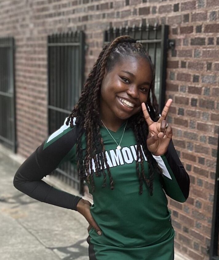 This photo is a full body shot of Janiya Thurman, a student at Gary Comer College Prep. She is a young Black woman with long braids pulled into a ponytail. In this photo, she is smiling and posing with a peace sign and wearing her green-and-white Gary Comer cheer team uniform.