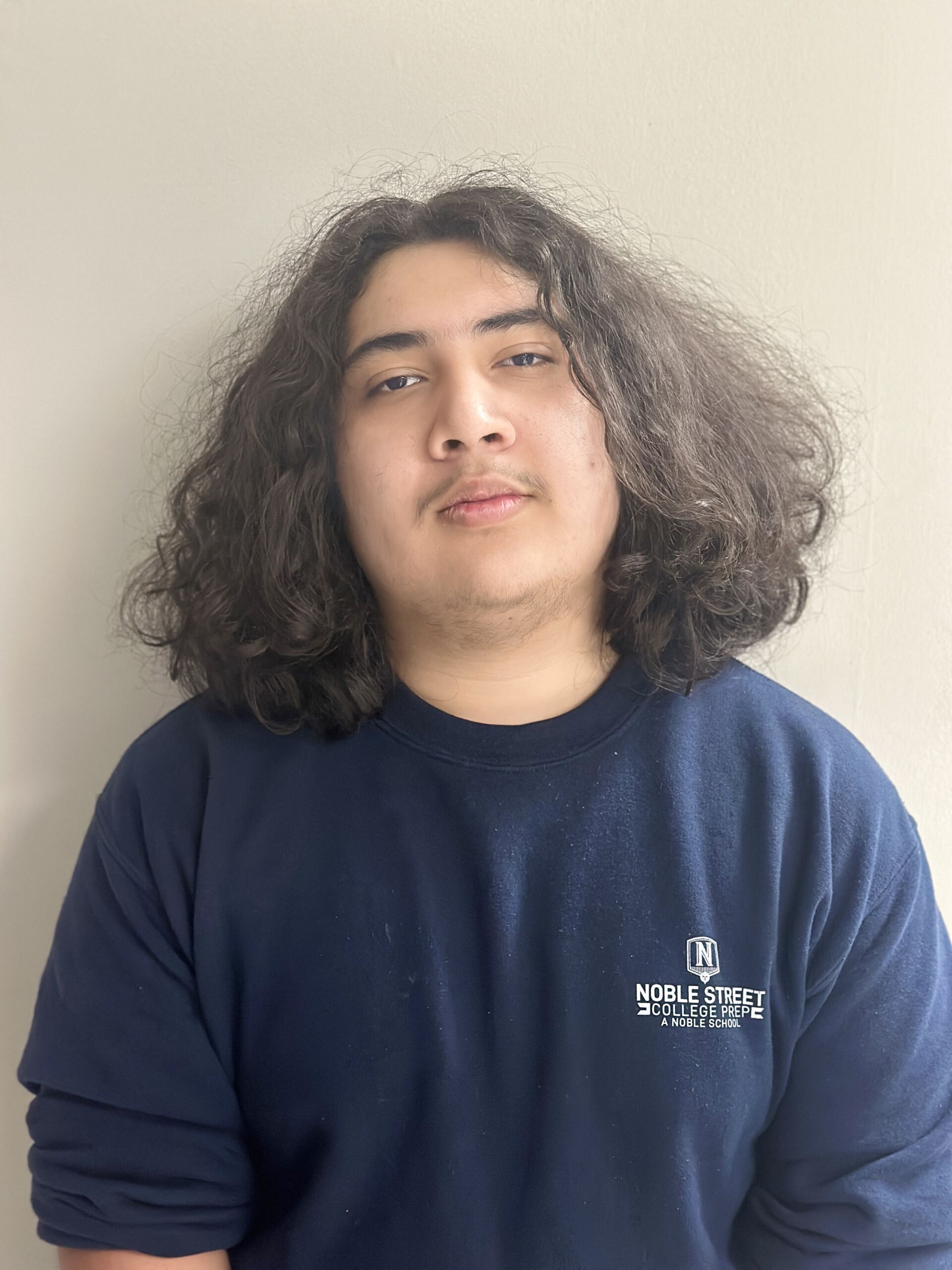 This photo shows a headshot of Miguel Carbajal, a student at Noble Street College Prep. He is a young Latine man with shoulder-length wavy hair. In this photo, he is wearing a dark blue sweatshirt with the Noble Street logo on it.
