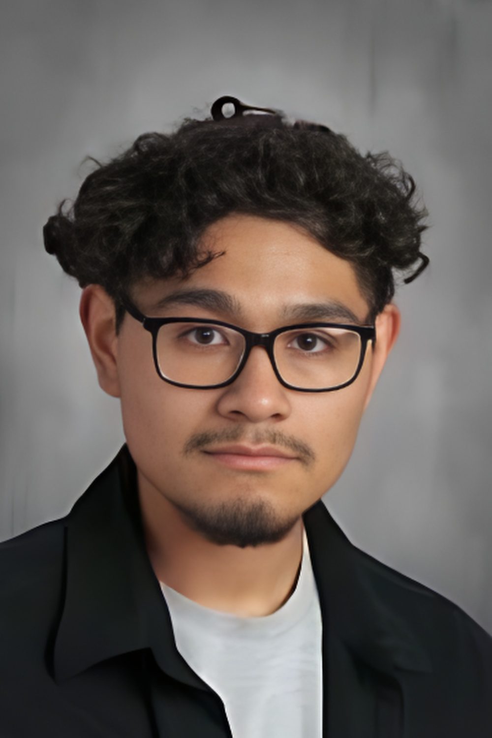 This photo is a headshot of Ernesto Sosa, a student at Mansueto High School. He is a young Latine man with short curly hair and glasses. In this photo, he is wearing a light gray t-shirt underneath a black button-up.