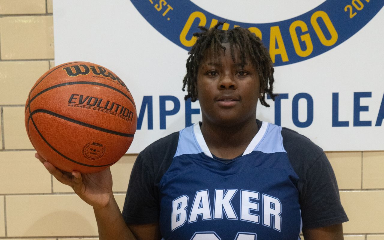 Photo shows Serenity Curry, a recent alum of Baker College Prep. She is a Black girl with short locs and is wearing her Baker basketball uniform. She is holding a basketball in her right hand.
