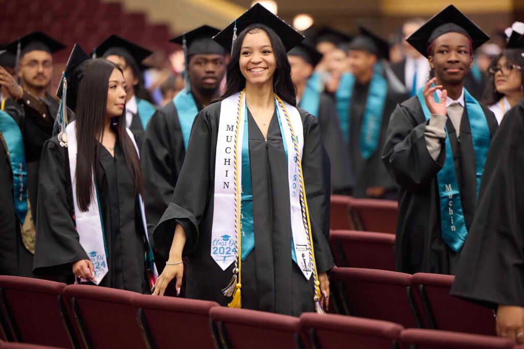 Image shows a smiling young woman who is graduating in Muchin College Prep's Class of 2024. She has her cap and robe on and is surrounded by her peers as they walk out of the auditorium.