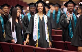 Image shows a smiling young woman who is graduating in Muchin College Prep's Class of 2024. She has her cap and robe on and is surrounded by her peers as they walk out of the auditorium.