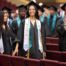 Image shows a smiling young woman who is graduating in Muchin College Prep's Class of 2024. She has her cap and robe on and is surrounded by her peers as they walk out of the auditorium.