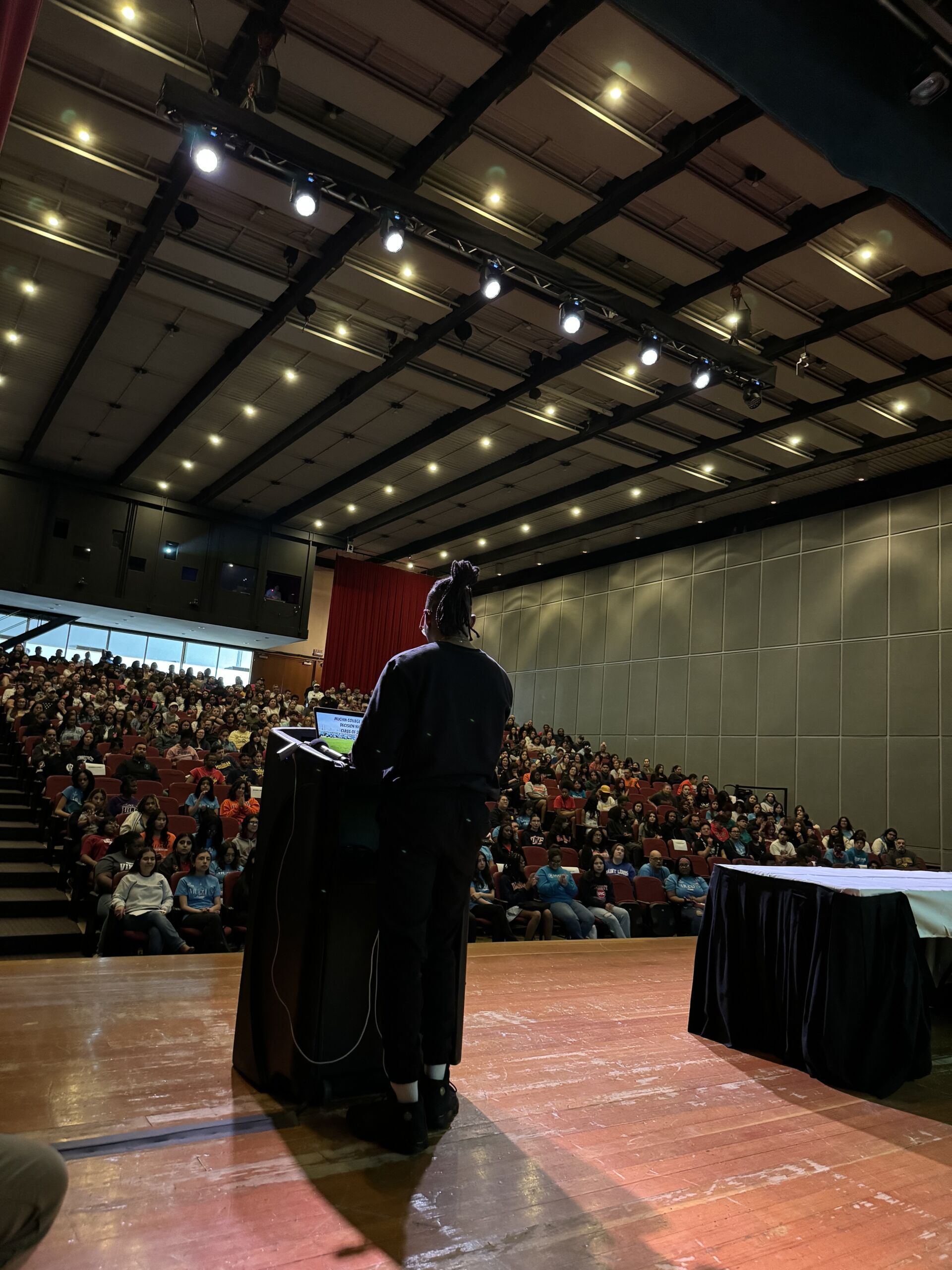Emmanuel Jackson, a college counselor at Muchin College Prep, stands in front of a podium on a stage. You can see the crowd of students in front of him in a large auditorium.