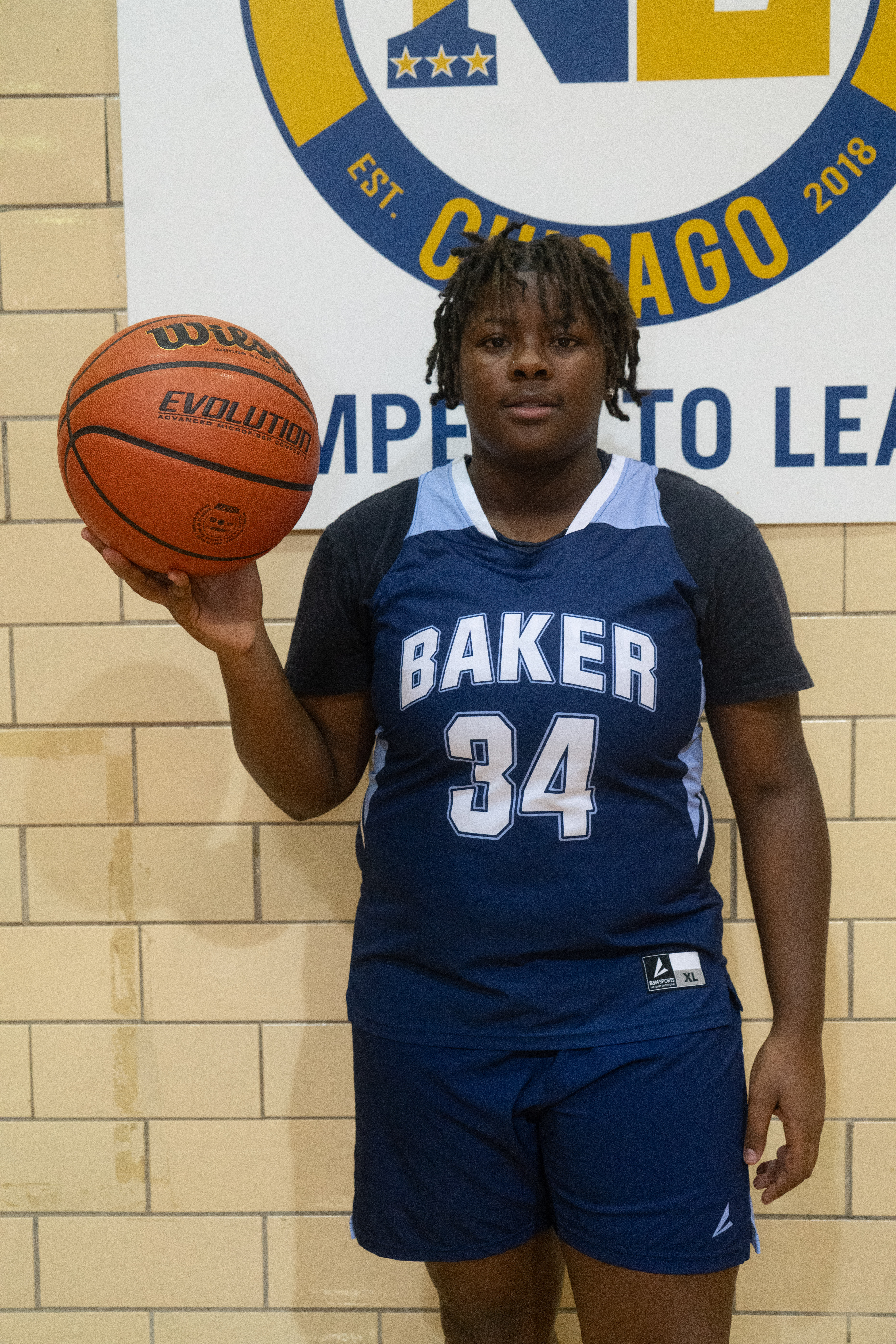 Photo shows Serenity Curry, a recent alum of Baker College Prep. She is a Black girl with short locs and is wearing her Baker basketball uniform with the number 34 on it. She is holding a basketball in her right hand.