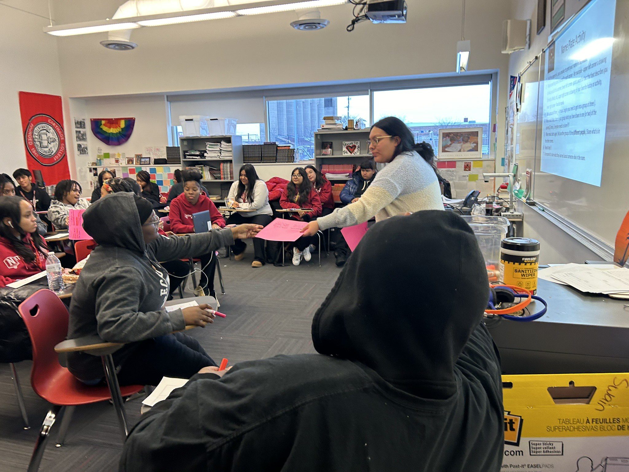 This photo shows a classroom at ITW David Speer Academy where many Black and Brown students are gathered in seats, working on an activity. You can see a teacher in the middle, handing a sheet of bright pink paper to one student at a desk. Behind the teacher is a presentation slide projected onto the whiteboard. The title of the slide is "Name Plate Activity". This is a meeting for Speer's Black and Brown Solidarity Club.