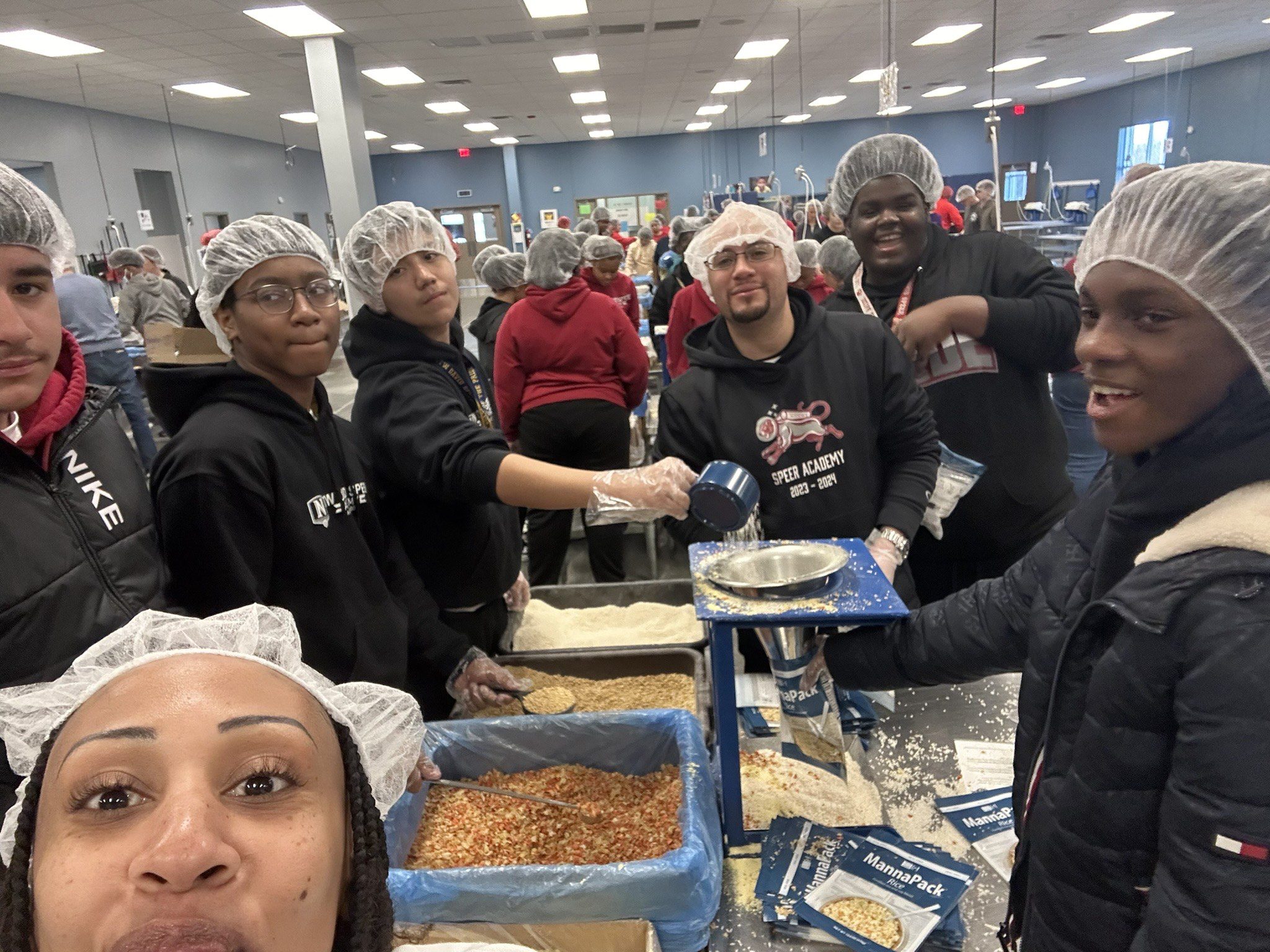 Photo shows a group selfie of ITW David Speer Academy students volunteering at a local kitchen for Feed My Starving Children. They are all wearing gloves and hair caps as they stand around a table with food stuffs on it that they are working on packaging.