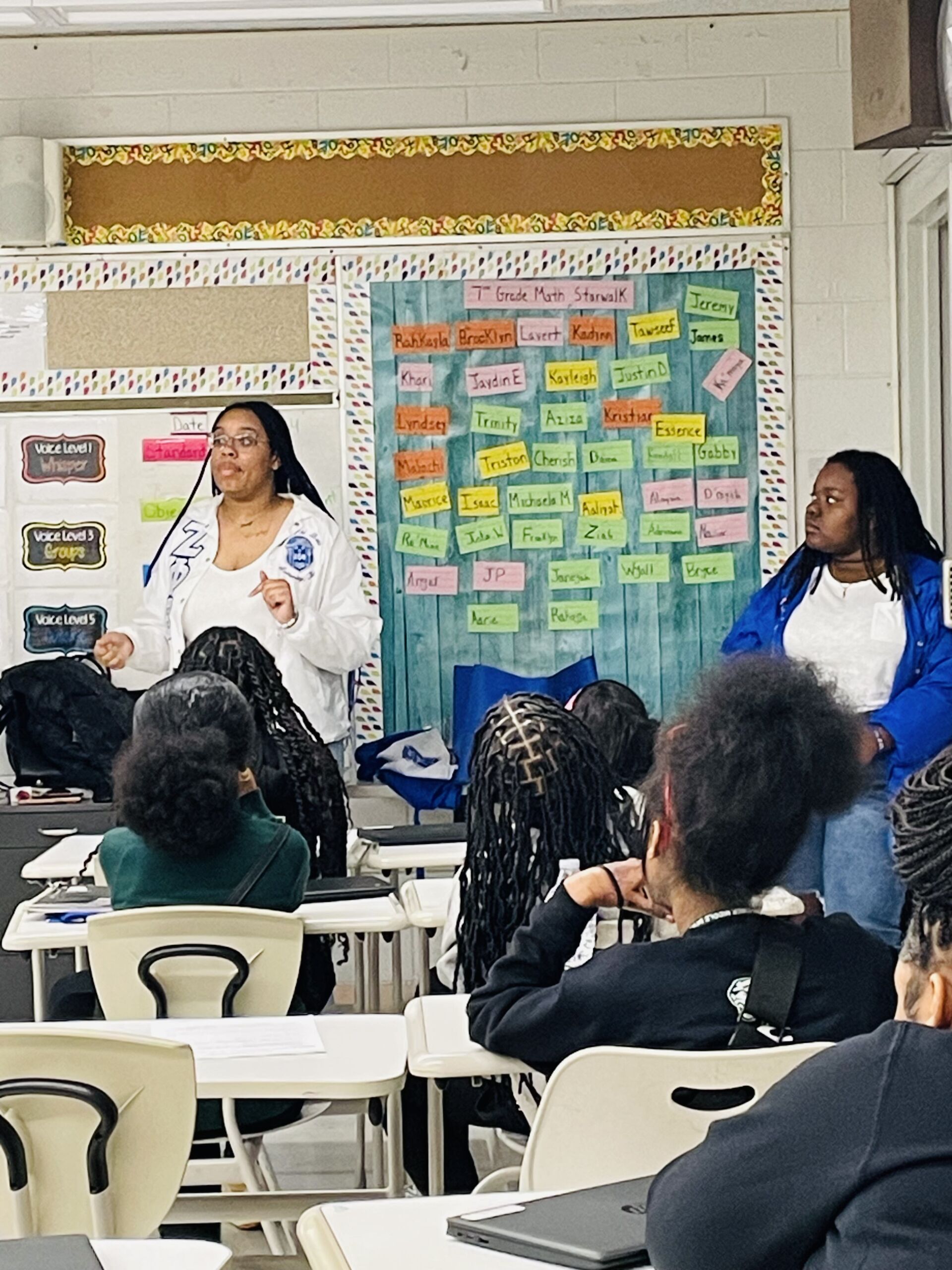 Two young Black women stand at the front of a middle school classroom. They are wearing blue and white Zeta Phi Beta sorority swag.