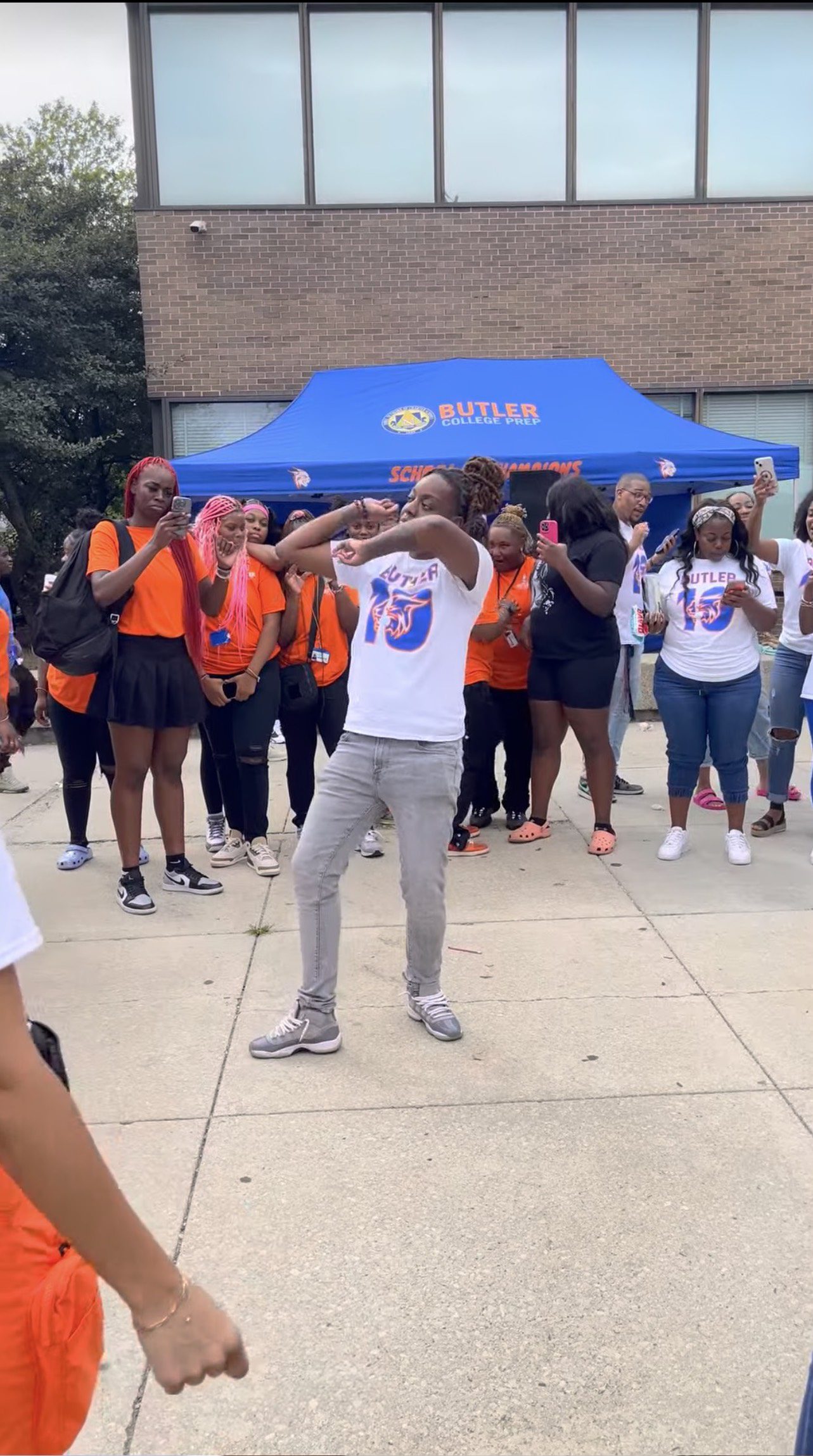 Benton dances outside, surrounded by Butler students and staff. She has her arms crossed in front of her face as she leans back. She's wearing a white Butler t-shirt and gray jeans and has her locs up in a bun.