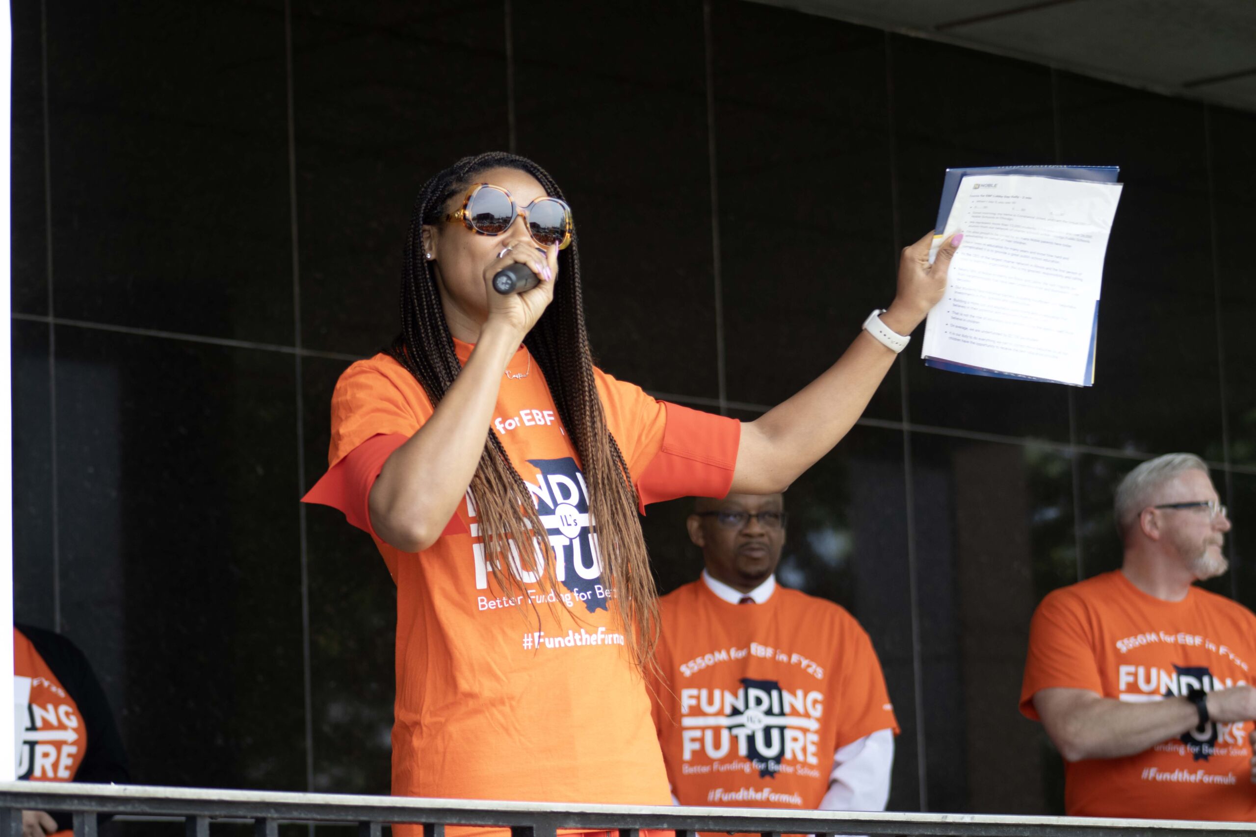 Jones gives a speech in front of a rally for Evidence-Based Funding. She is holding a mic in one hand and a paper in the other. She is making a gesture like she is amping up the crowd as she speaks.