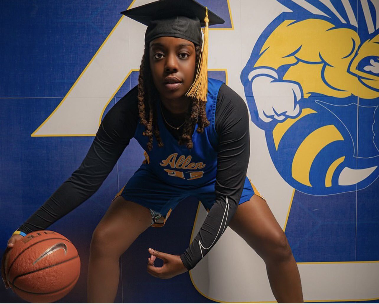 Benton is crouching and dribbling a basketball in front of a wall decal with the Allen University mascot and colors. She wears her Allen University basketball uniform and has a graduation cap on top of her shoulder-length brown locs.