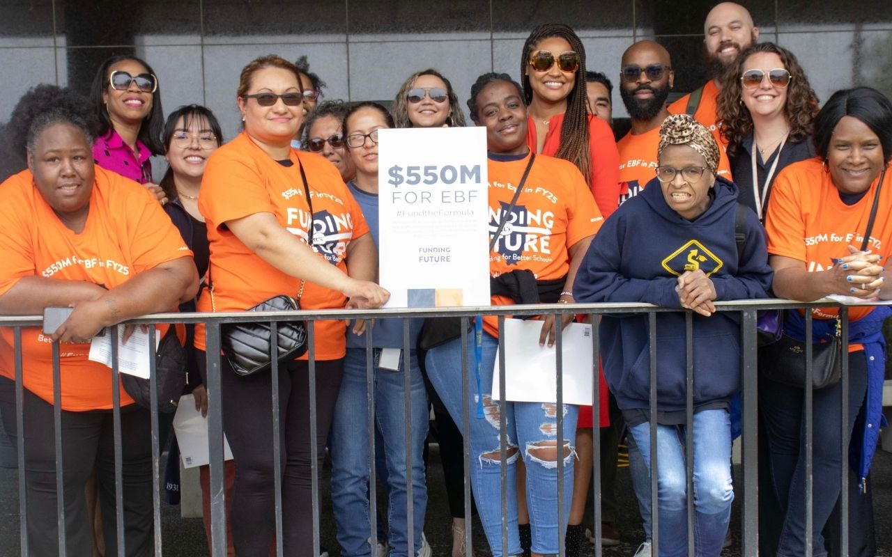 A contingent of Noble Schools' parents, alumni, and staff pose with posters calling for Evidence-Based Funding in Illinois. They are all wearing bright orange shirts that say Funding Illinois Future on it.