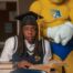 Illie Benton sits at a desk with books in front of her and the Allen University mascot behind her. She is wearing a graduation cap, a long-sleeve button-up and a sweater vest.