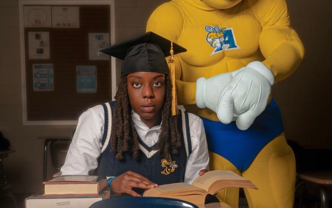 Illie Benton sits at a desk with books in front of her and the Allen University mascot behind her. She is wearing a graduation cap, a long-sleeve button-up and a sweater vest.
