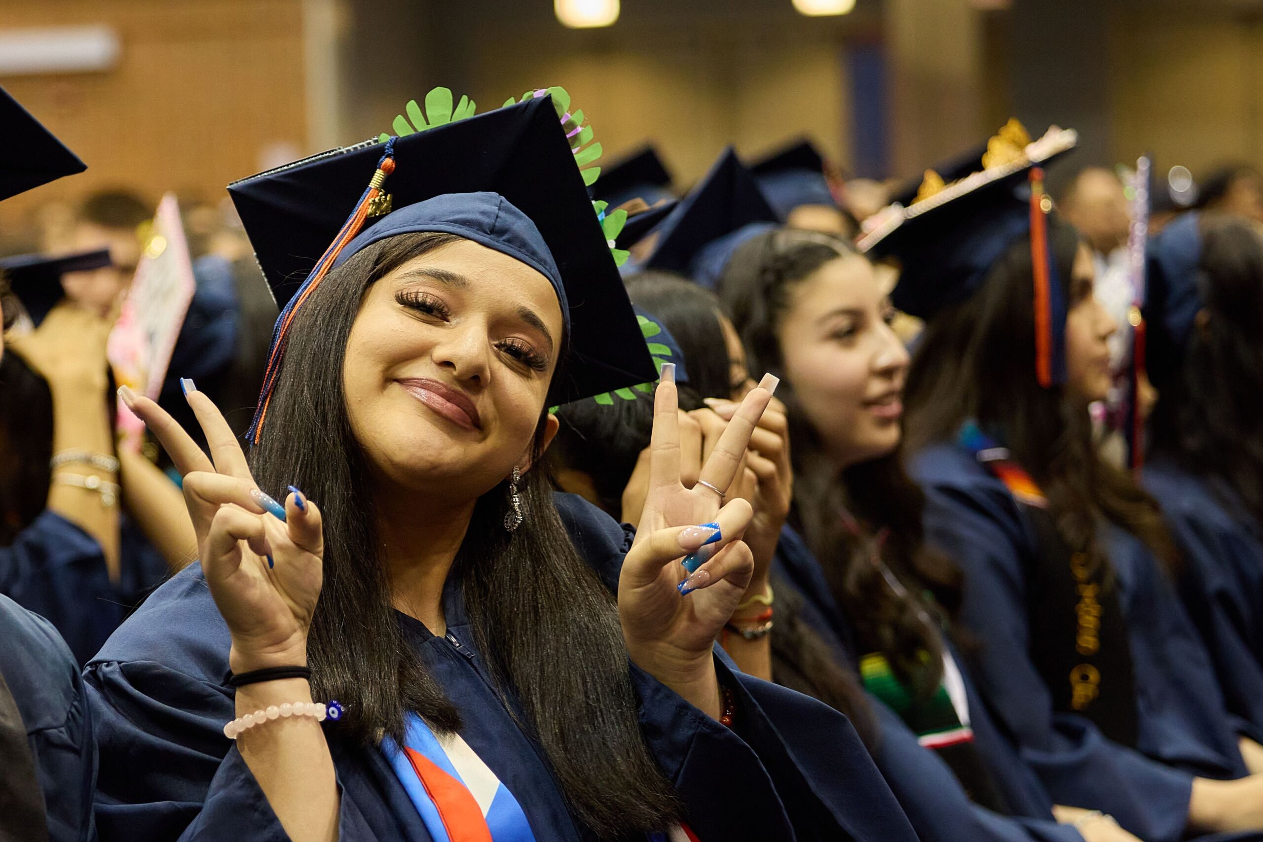 A young Latinx student in a cap and gown smiles and puts up two peace signs at their graduation.