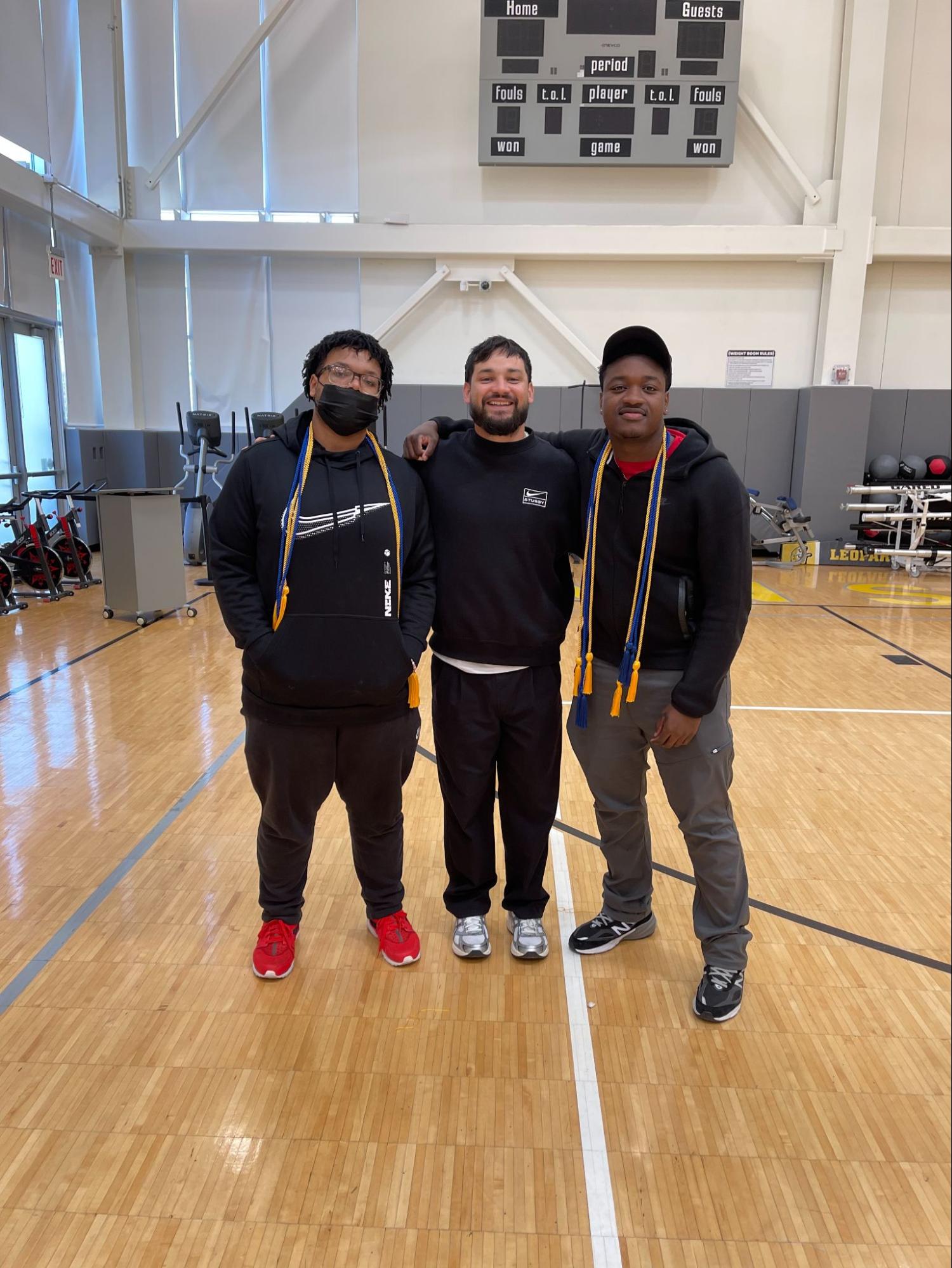 Walter and Deandre stand with their basketball coach, Jose Arteaga, in the Mansueto High School gym. The two boys are wearing graduation cords around their neck.