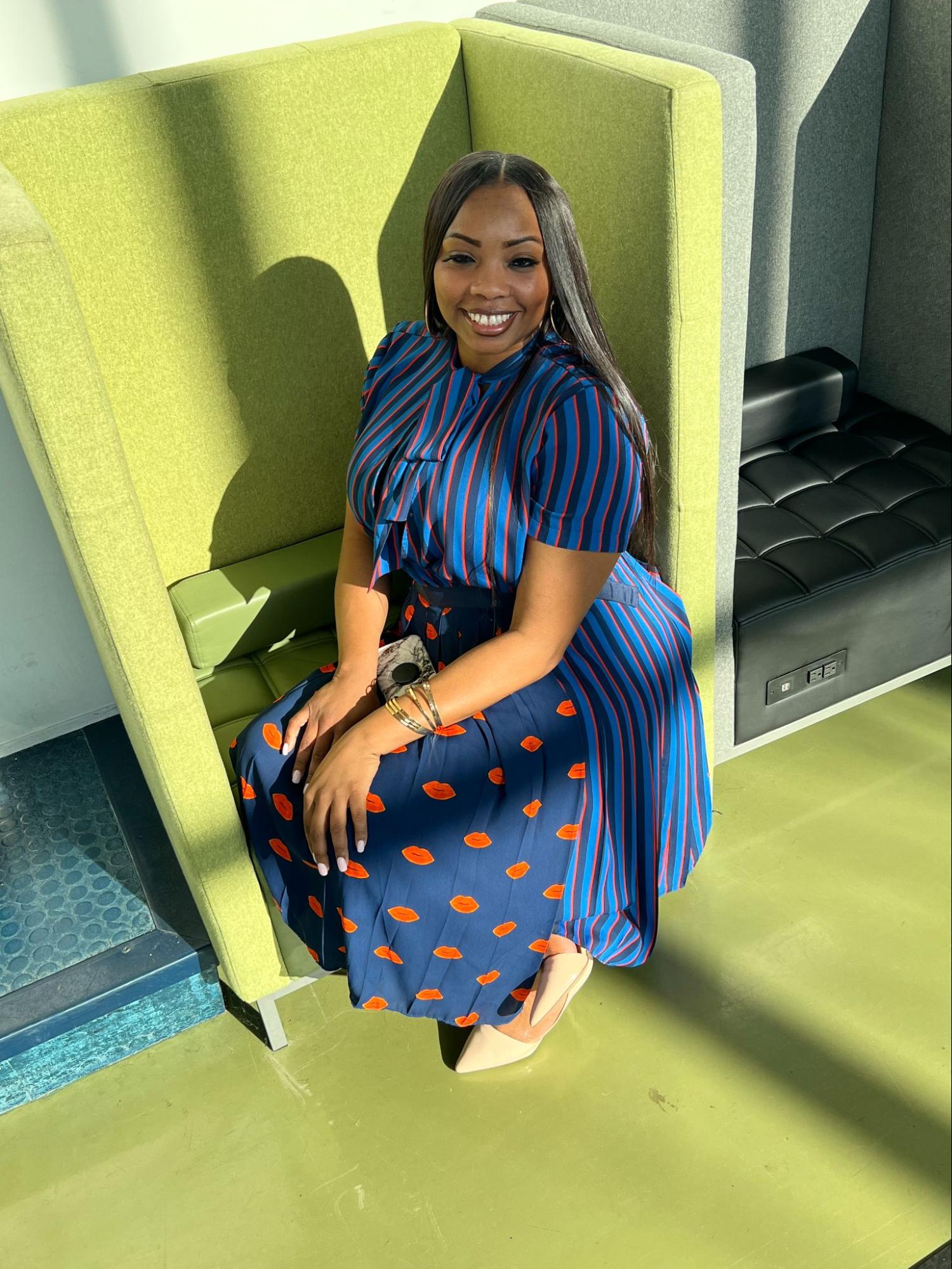 Chloe Hassell sits in a light green chair with high walls like a cubicle. She has medium-light brown skin and long straight dark brown hair. She is wearing a colorful dress with stripes on the top and back and lip mark graphics on the front bottom half of the skirt. It is navy, light blue, and red. She is smiling.