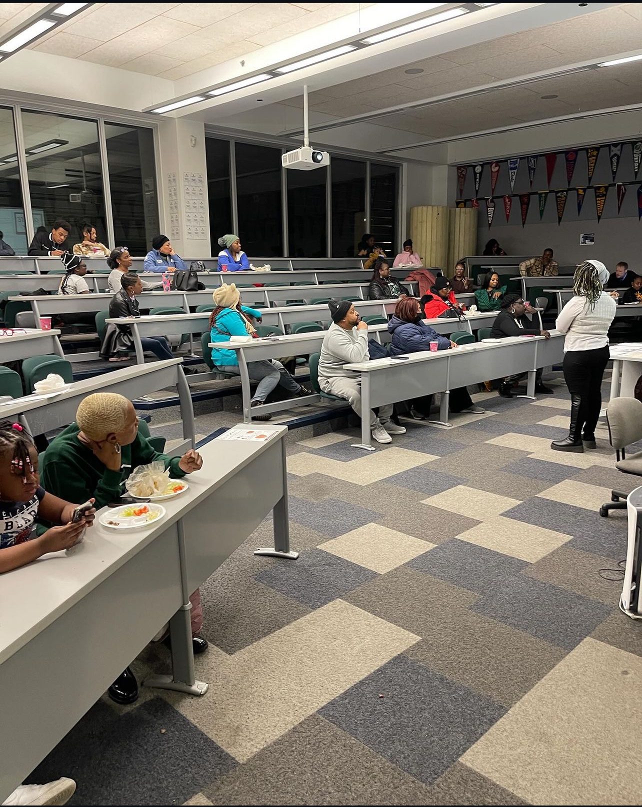 A group of Gary Comer College Prep parents and their kids sit at long rows of tiered desks in a lecture room space. They are listening to a staff member talking at the front of the room.