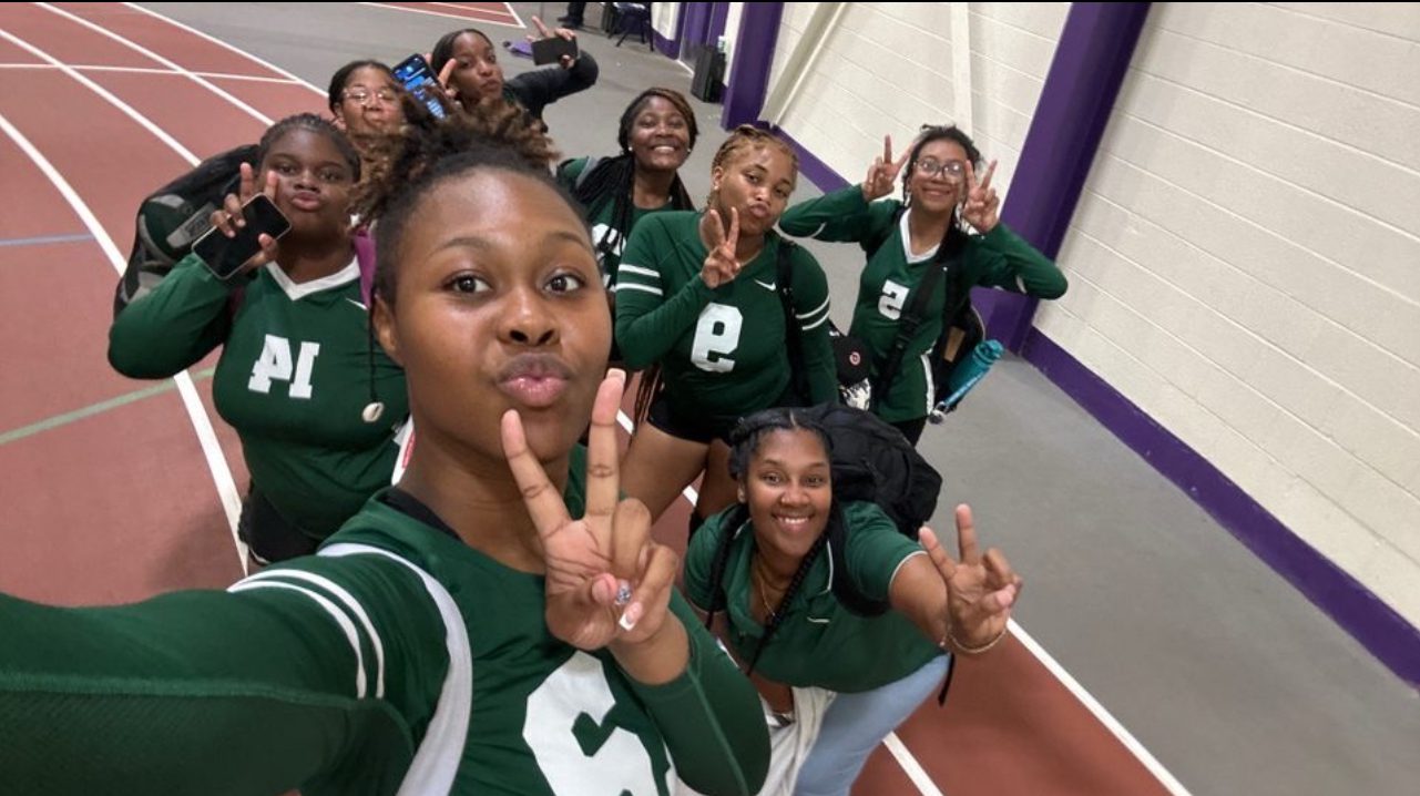 A selfie of the Comer girls volleyball team after the game. They are all in their long-sleeved, dark green uniforms and throwing up peace signs. In the back of the group, you can see Laramie Bruce, GCCP parent and coach, also throwing up a peace sign.