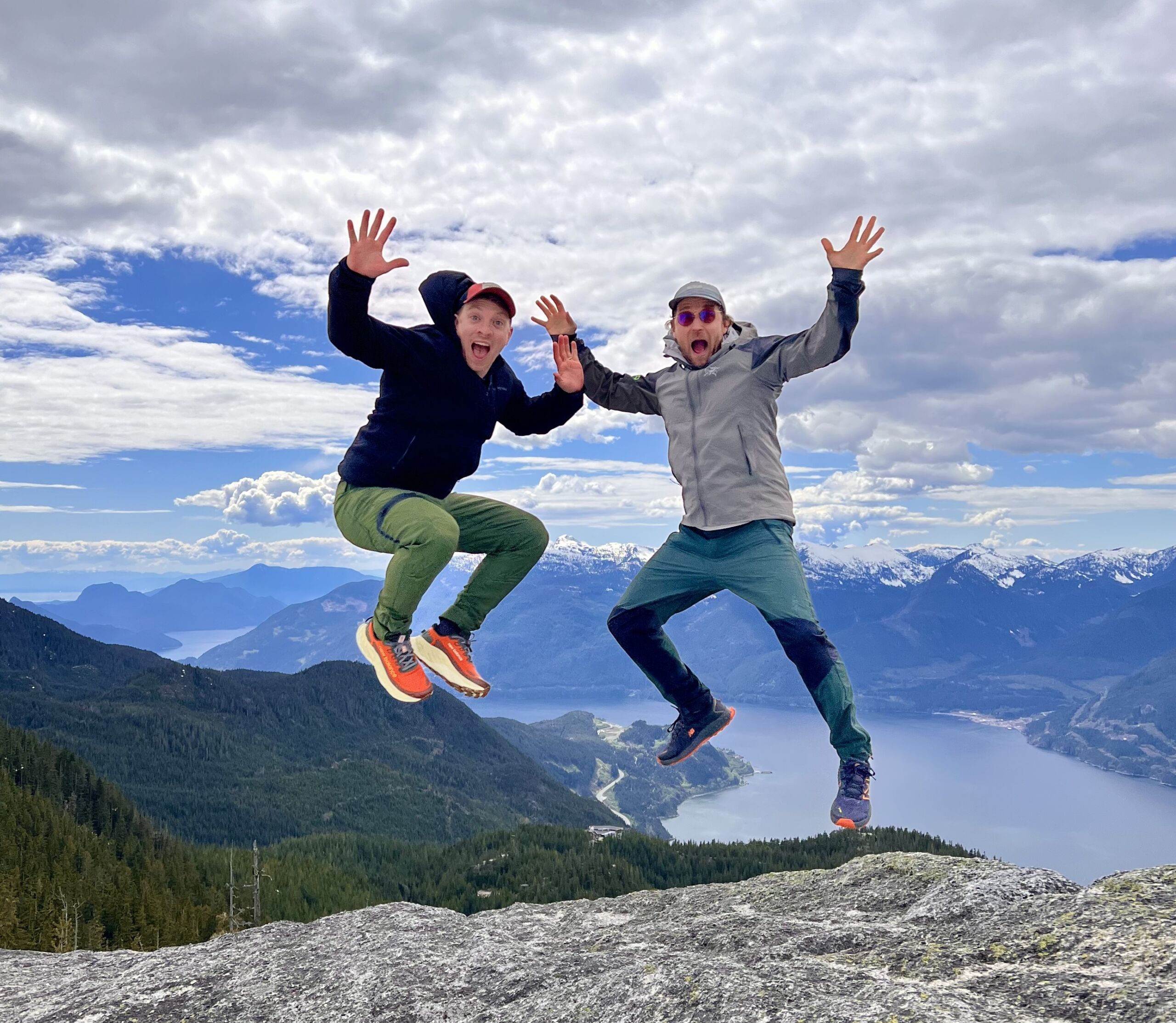 Gunty and a friend or family member jumping into the air in front of a gorgeous mountain landscape. They are smiling wide with their hands splayed open, giving a celebratory and silly look.