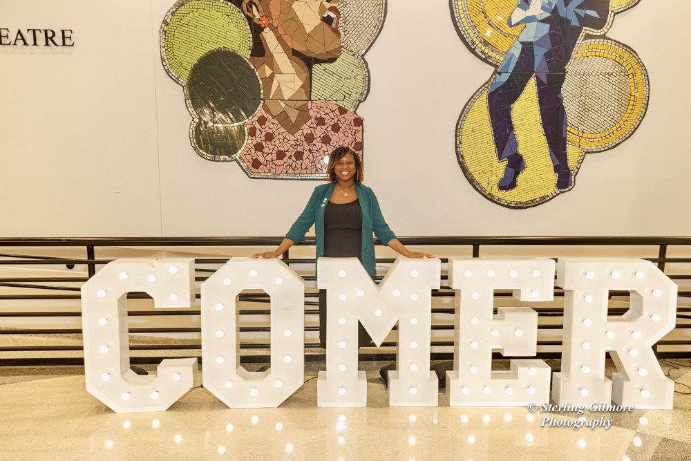 Hemingway standing and smiling behind a big sculpture sign that says "Comer"