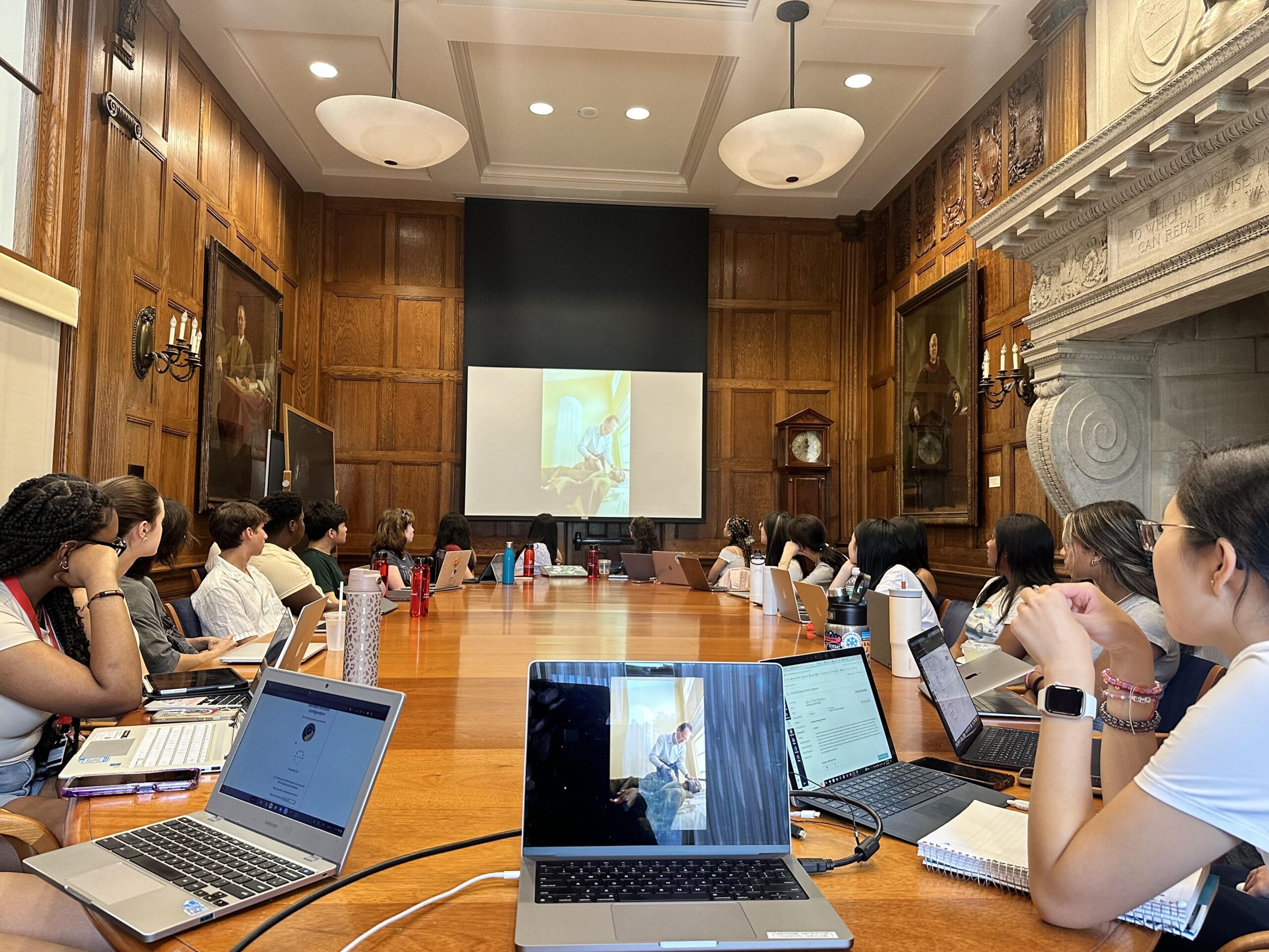 High school students from all over sit at a long wooden table in a lecture hall at Harvard University. They all have their laptops out and are paying attention to the presentation at the front of the room.