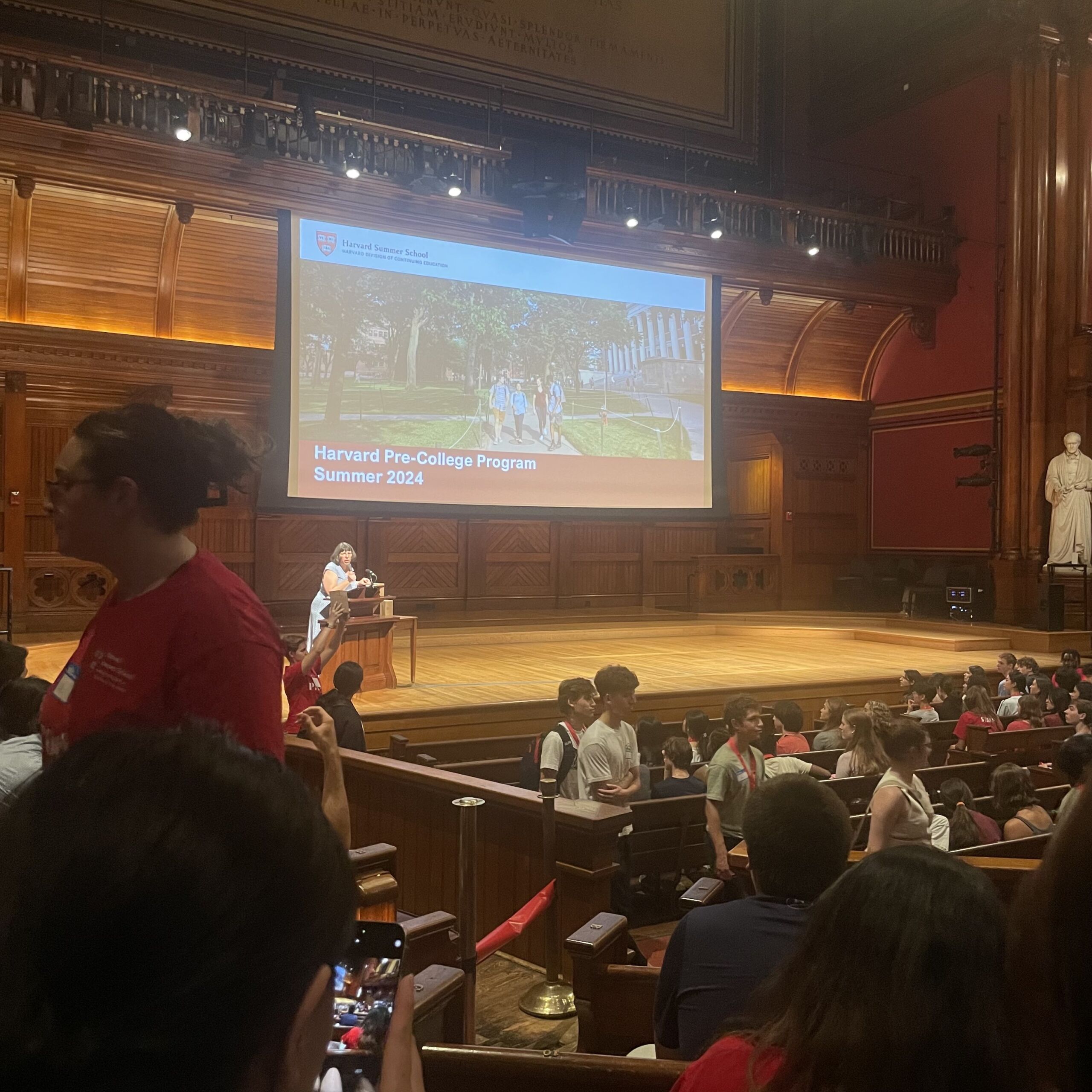 A crowd of high school students from all over are getting settled in an auditorium at Harvard University. The PowerPoint on the projector screen above the stage shows a photo of Harvard's campus with the words "Harvard Pre-College Program Summer 2024". A woman is speaking at the podium on the stage.