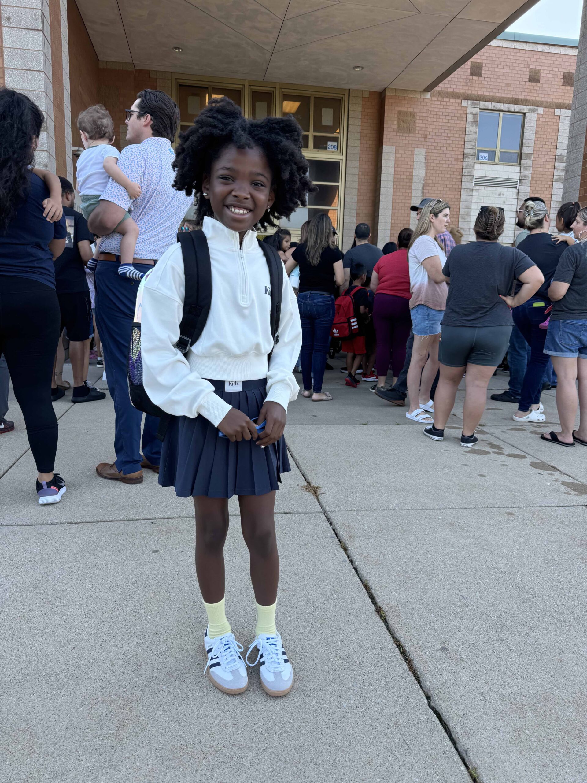 Cleaves' young child outside a building and amongst a crowd of people. She looks ready for school with a uniform and backpack on.