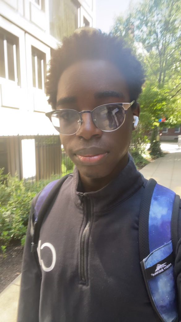 A selfie of Emmanuel, a student at Baker College Prep, while he is outside. He has short dark brown hair, dark brown skin, and is wearing glasses, a black sweatshirt, and a blue backpack.