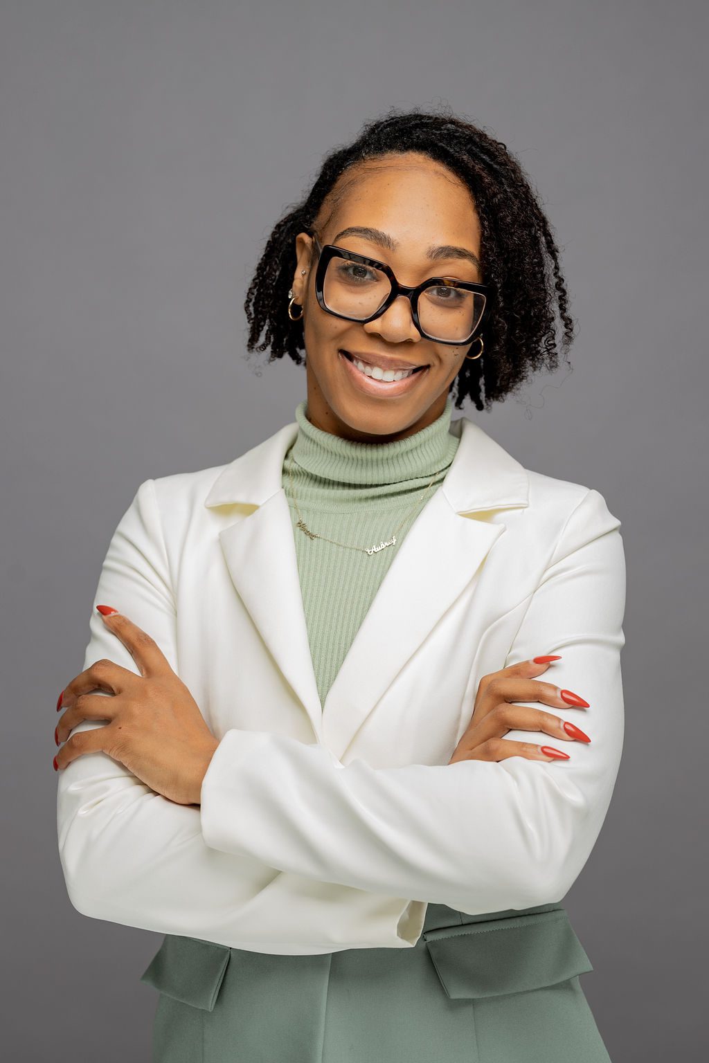 Headshot of Apiphany Asberry. She is woman with medium-light brown skin and short black locs that go down to mid-cheek. She is smiling and wearing glasses and a white and green blazer over a light green turtleneck.