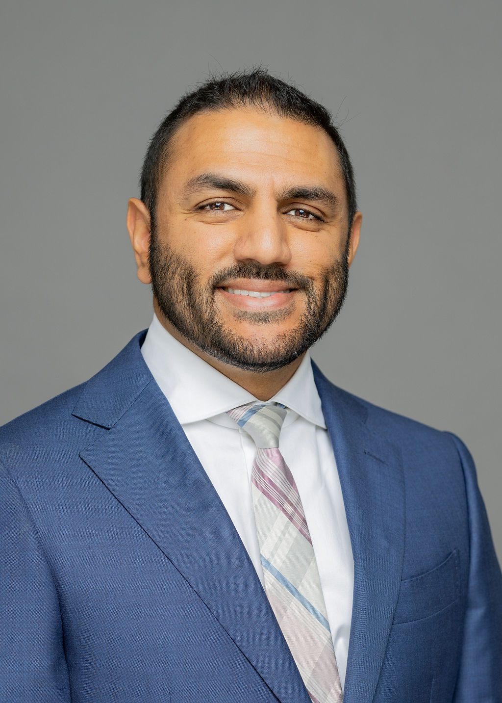 Headshot of Amit Khatkhate. He is a man with light brown skin, short black hair in a buzz cut, and a short beard. He is smiling and wearing a blue suit with a light red, blue and white tie.