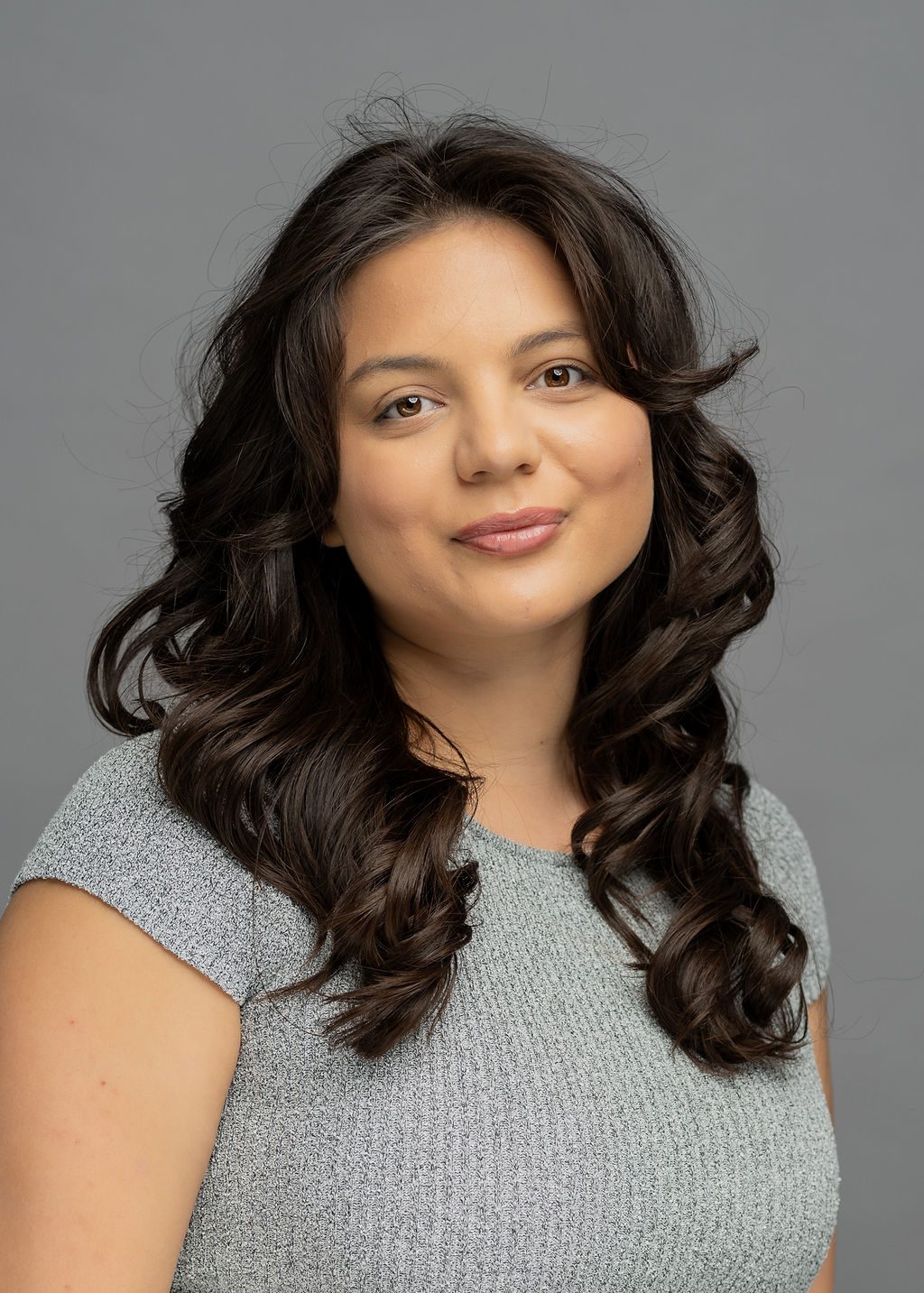 Headshot of Brenna Rivas. She is a woman with light brown skin and long wavy black hair. She is smiling and wearing a gray blouse.