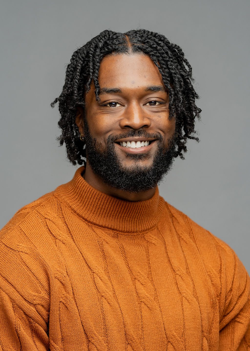 Headshot of Chantel Jones. He is a man with dark brown skin, chin-length black braids, and a short black beard. He is smiling and wearing an orange turtleneck.